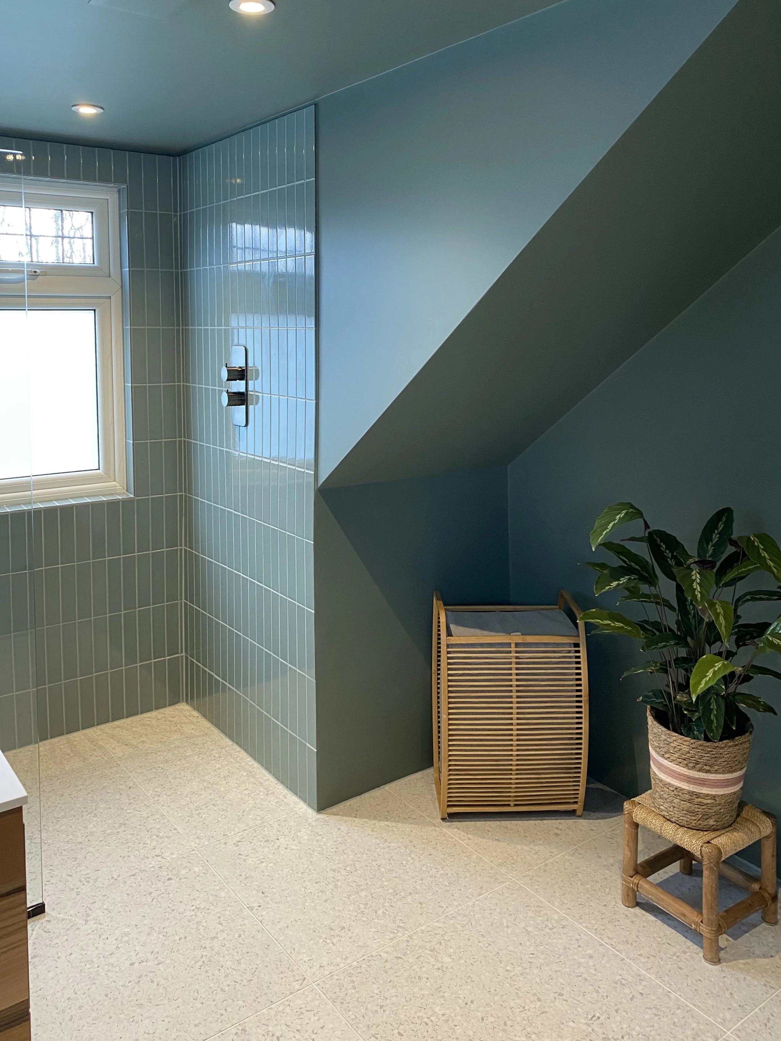 Compact bathroom addition under the eaves with colour-drenched warm blue/green walls, terrazzo flooring and vertical tiles, brushed black fixtures, and bespoke walnut vanity with Corian top.