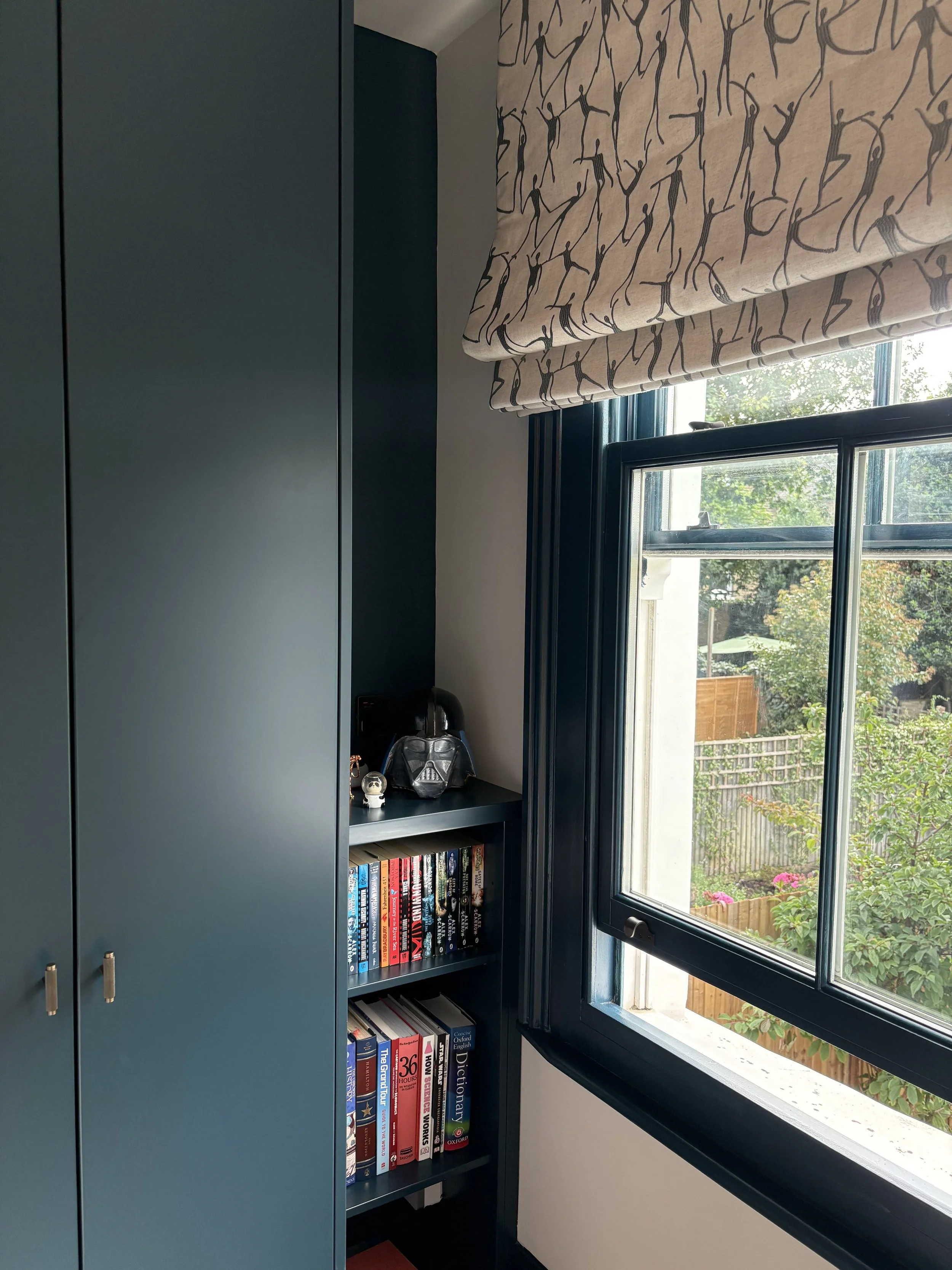 Bespoke dark blue fitted joinery - wardrobe and bookshelves for optimal storage. Window frame painted in dark blue for a bold accent and bespoke roman blinds with choreography theme for added fun and poesy.