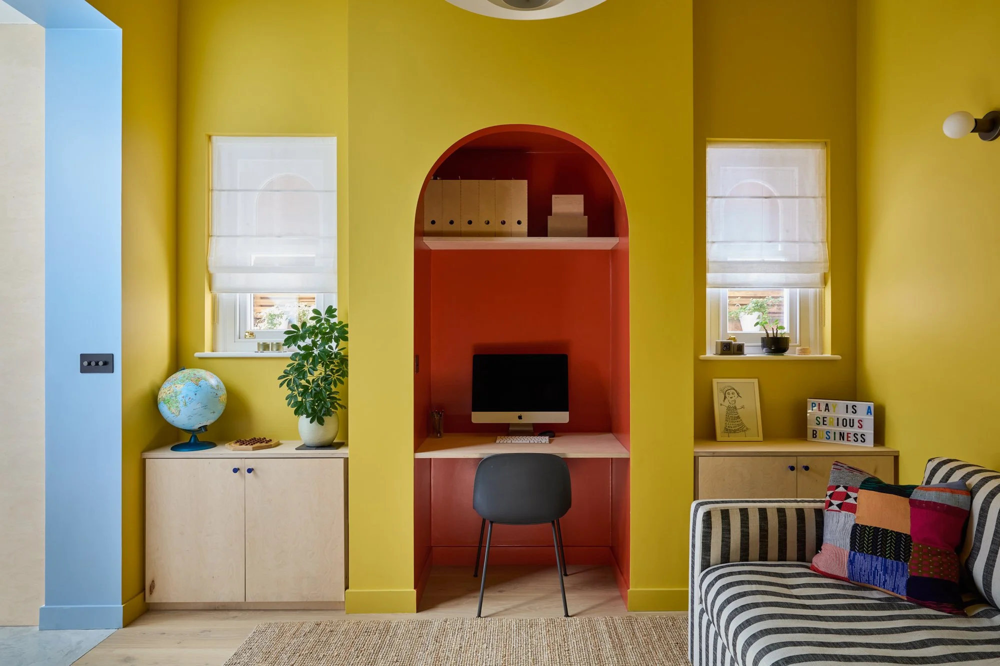 Vibrant playroom and family room with yellow, red, and blue accents. Features a chimney breast desk nook, bespoke birch plywood joinery, PH5 pendant, stripy sofa, jute rug, and decorative plants for a playful yet functional space.