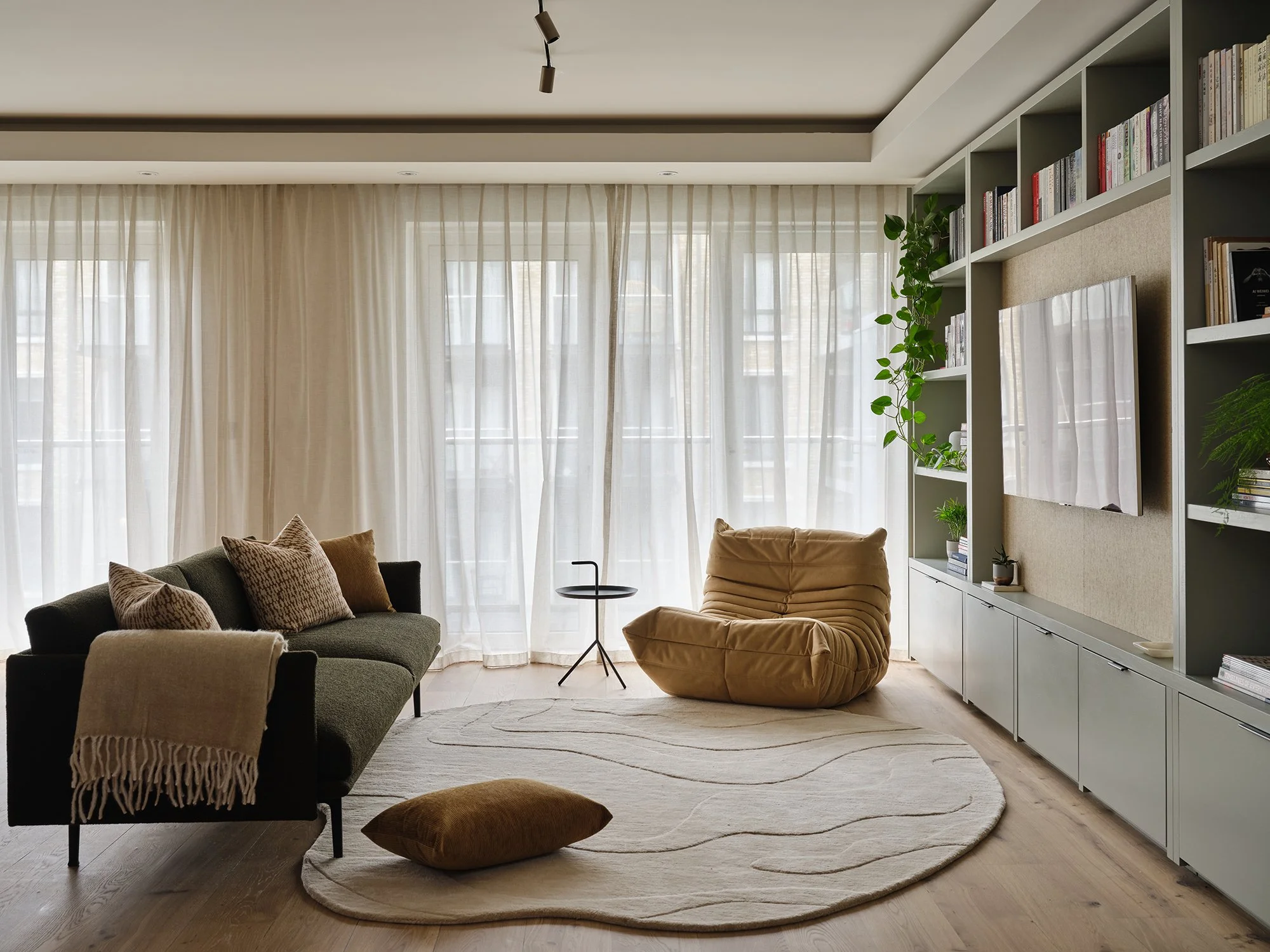 Contemporary living room in a Fulham Reach apartment with bespoke shelving, soft neutral textiles and full-height sheer curtains. Including iconic Togo chair. 