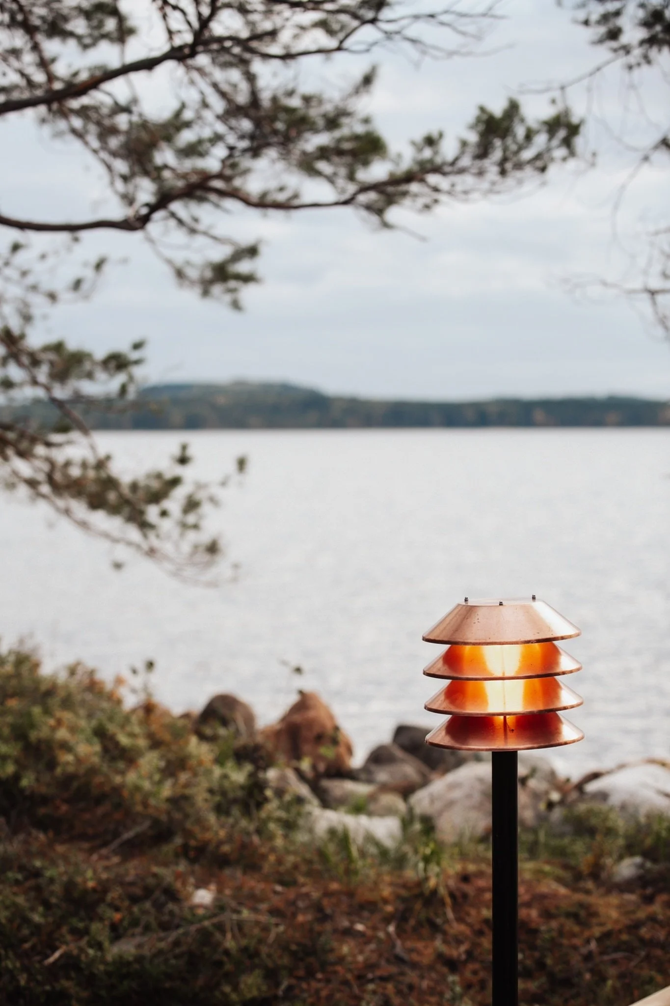Copper-finished outdoor lamp near a rocky lakeshore, framed by Finnish nature and soft water views in a peaceful, private setting.
