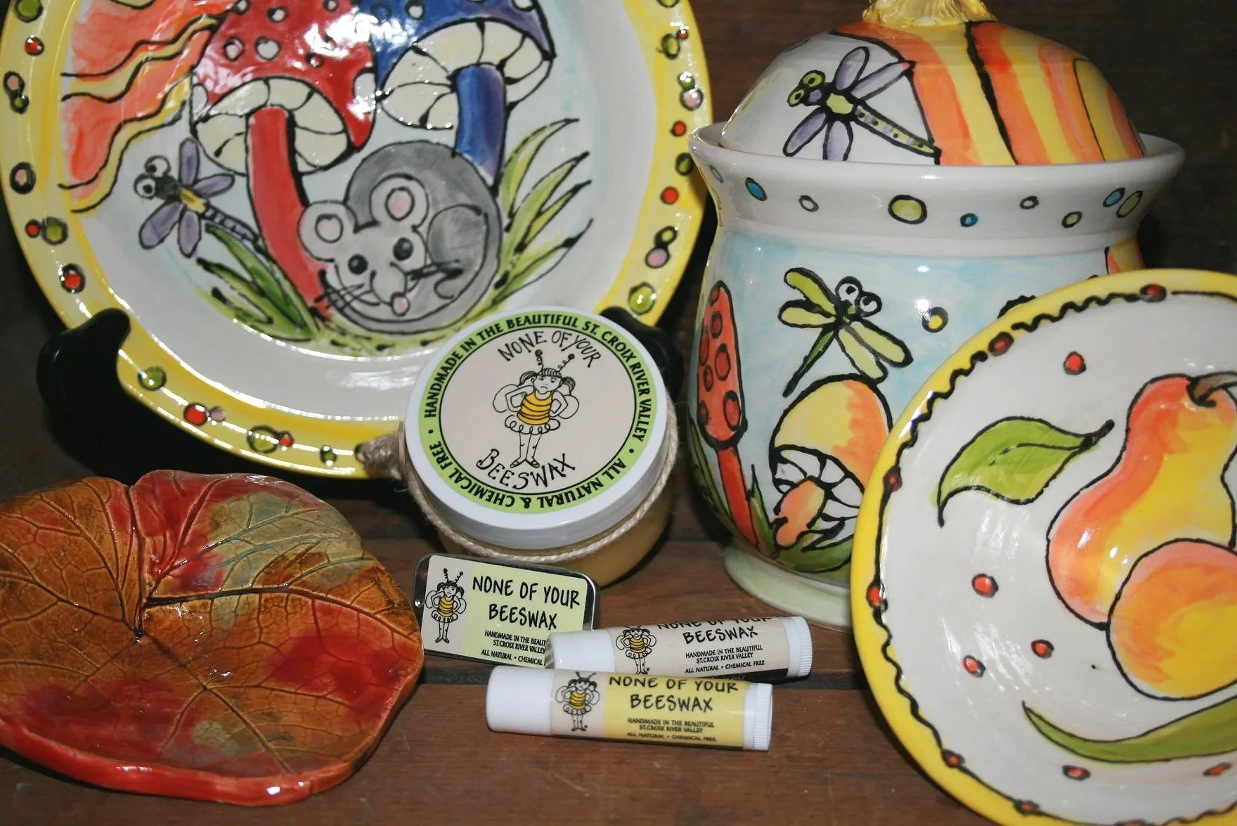 Colorful ceramic dishware with hand-painted designs of fruits, mushrooms, and insects, along with beeswax products and a decorative leaf-shaped dish.