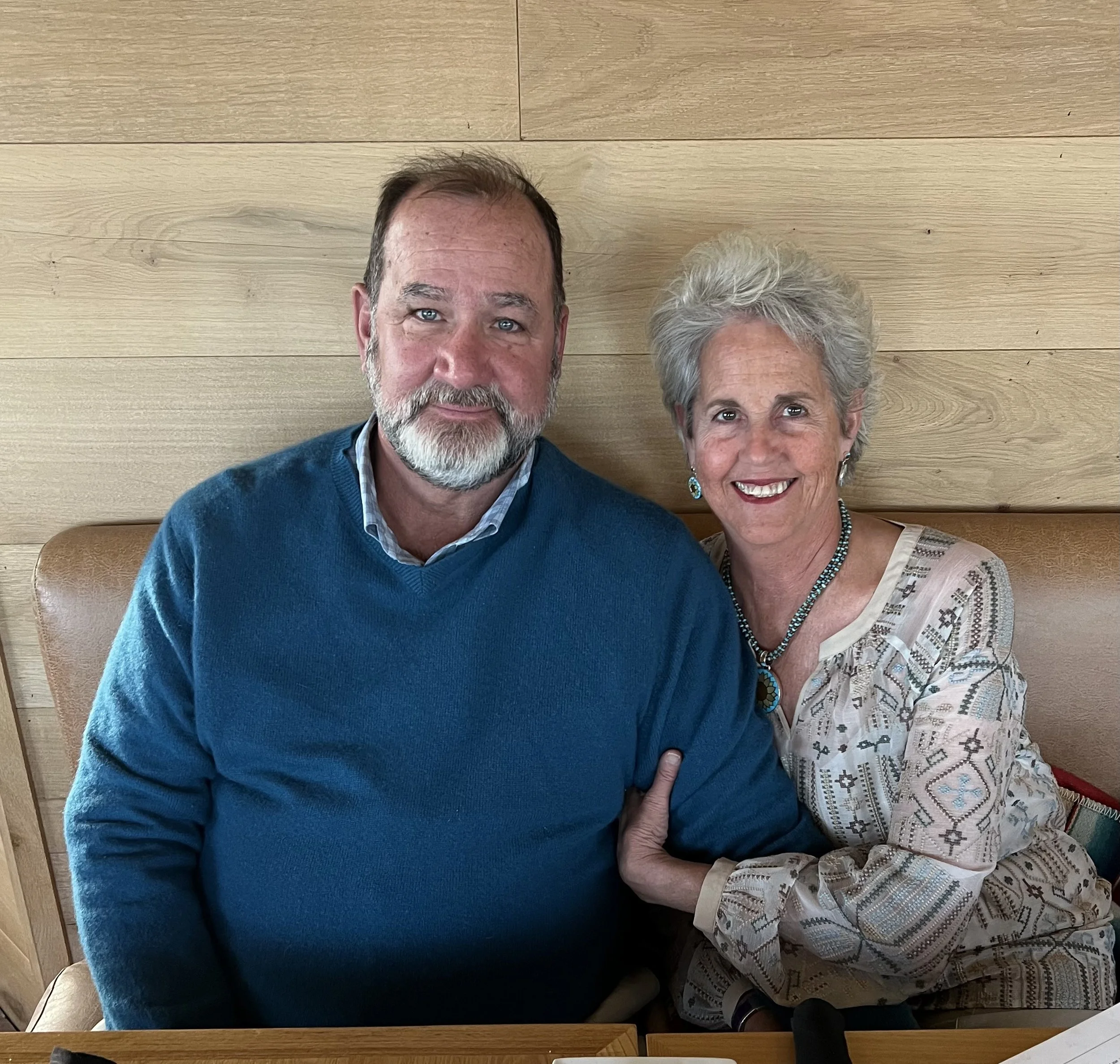 A smiling elderly woman with short gray hair and a beaded necklace sitting next to a man with a beard wearing a blue sweater, both seated at a table with a light wood wall background.