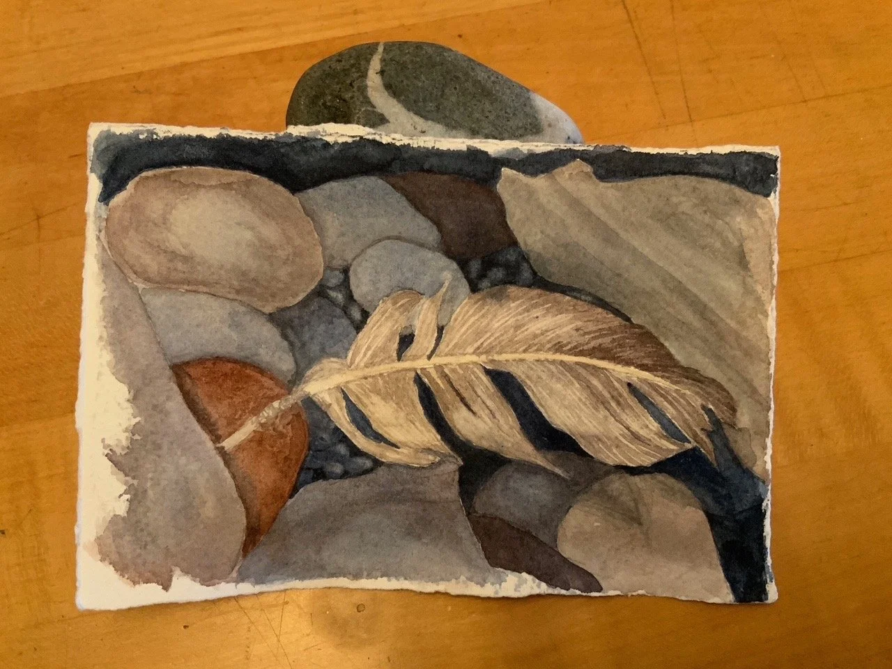 Watercolor painting of pebbles and a dried leaf on a wooden surface.