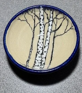 A round ceramic dish with a blue rim, featuring a painted scene of three white birch trees with black branches against a beige background.
