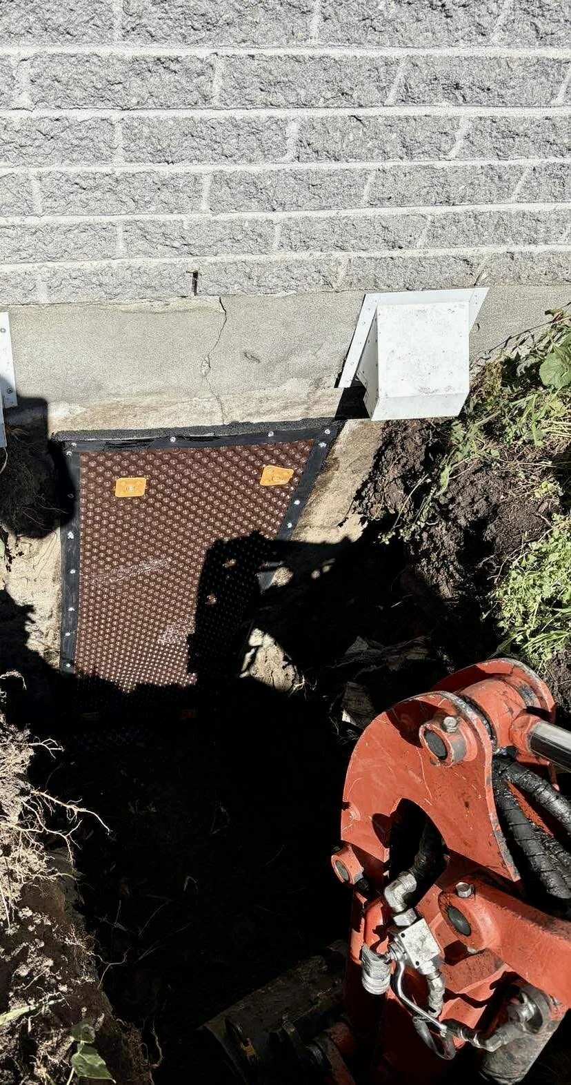 Travée d'accès souterraine en cours d'installation, avec une porte d'accès en métal, une boîte électrique blanche et un équipement hydraulique rouge, à proximité d'une maison avec un mur en briques.