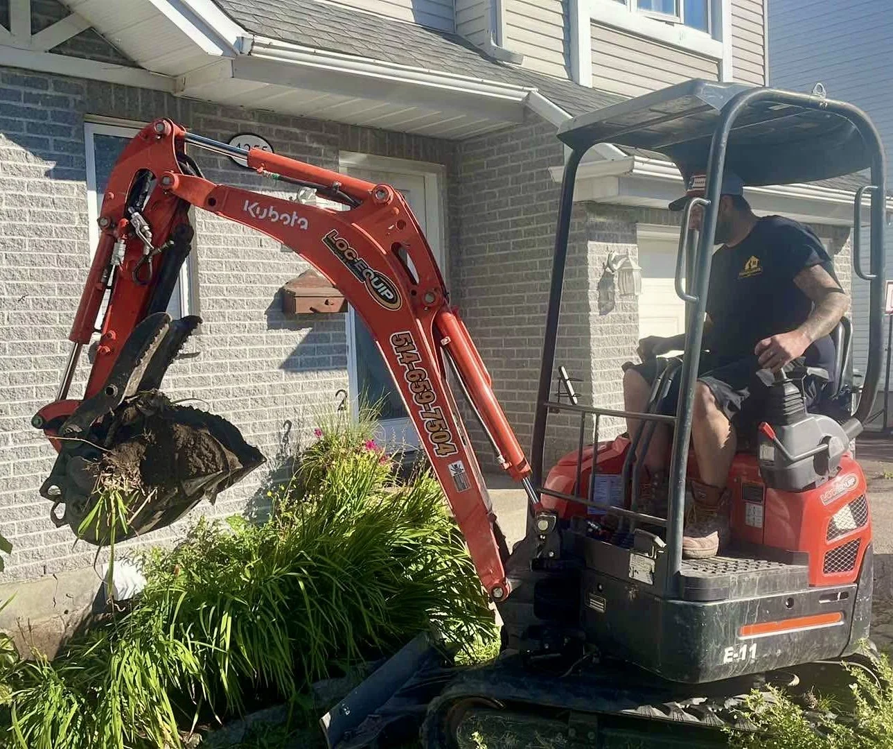 Un homme conduisant une pelleteuse rouge en train de déterrer une plante dans un jardin devant une maison à brick. L'homme porte un t-shirt noir, un casque de chantier et des chaussures de travail.