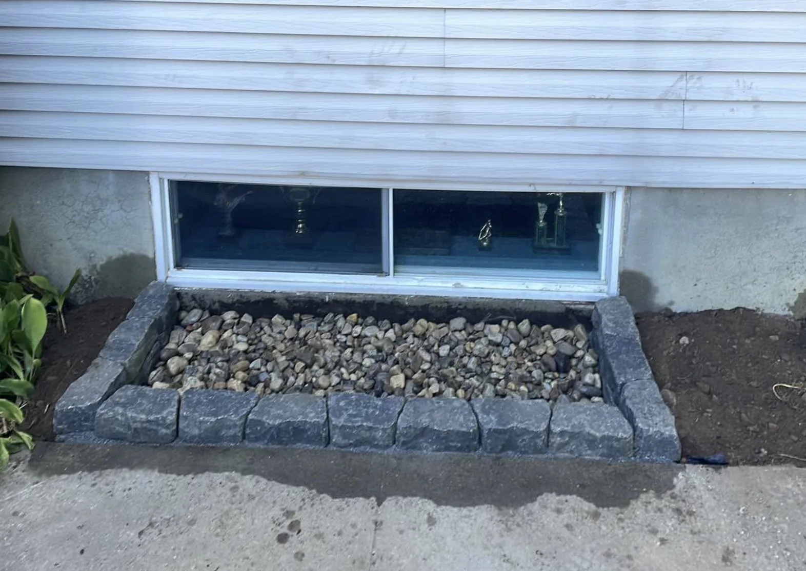 Fenêtre de sous-sol avec une frontière en pierre et des pierres au sol, entourée de terre et de végétation.