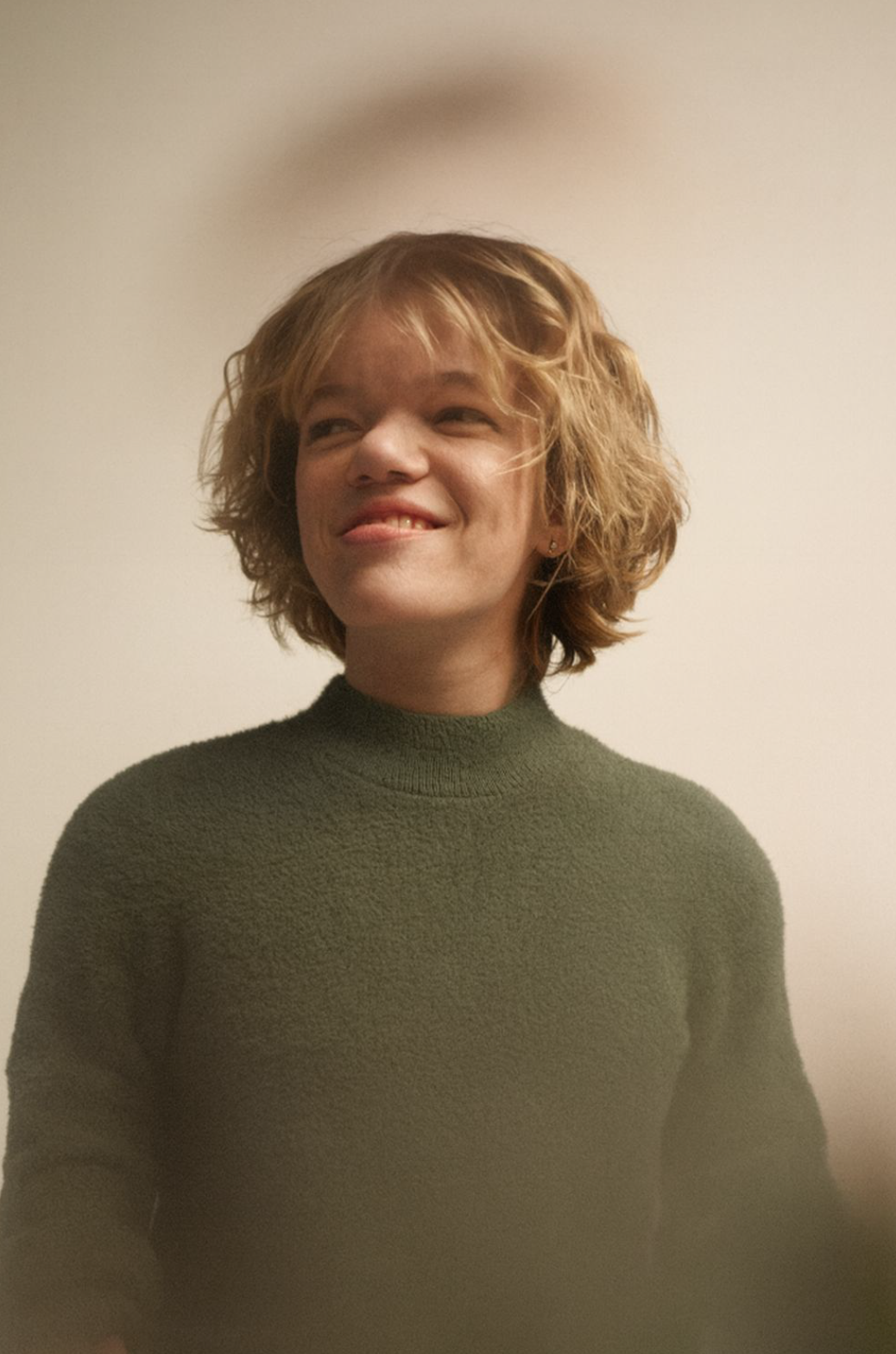 Joven mujer con cabello rizado rubio, sonrisa ligeramente torcida, usando suéter verde y ecléctico, fondo neutro.