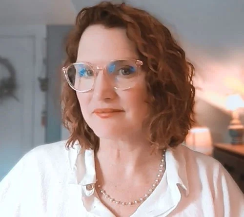 A woman with wavy red hair wearing glasses and a white shirt, in an indoor setting with a blurred background.