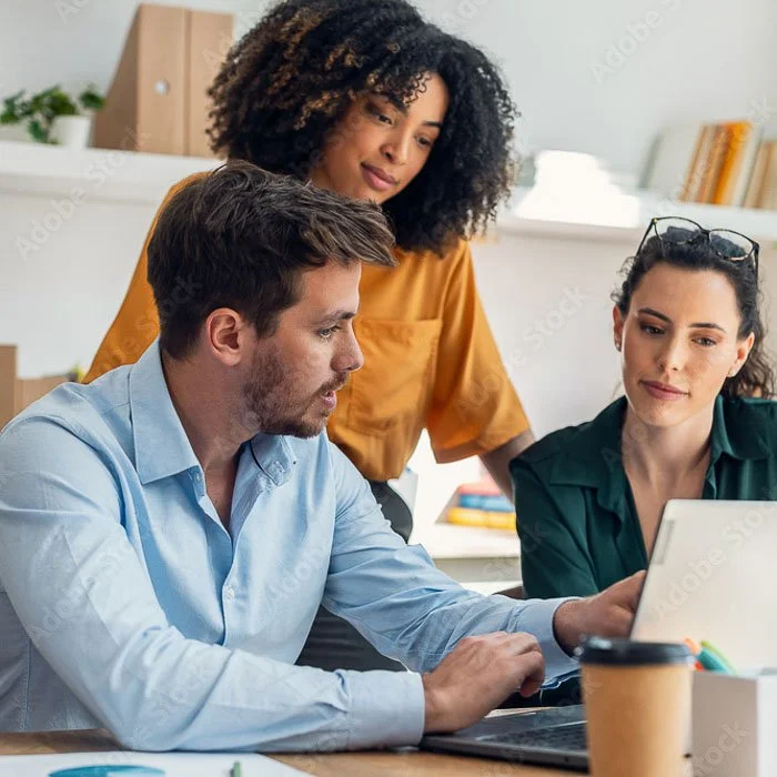Three colleagues working together around a laptop in an office setting, discussing something on the screen.