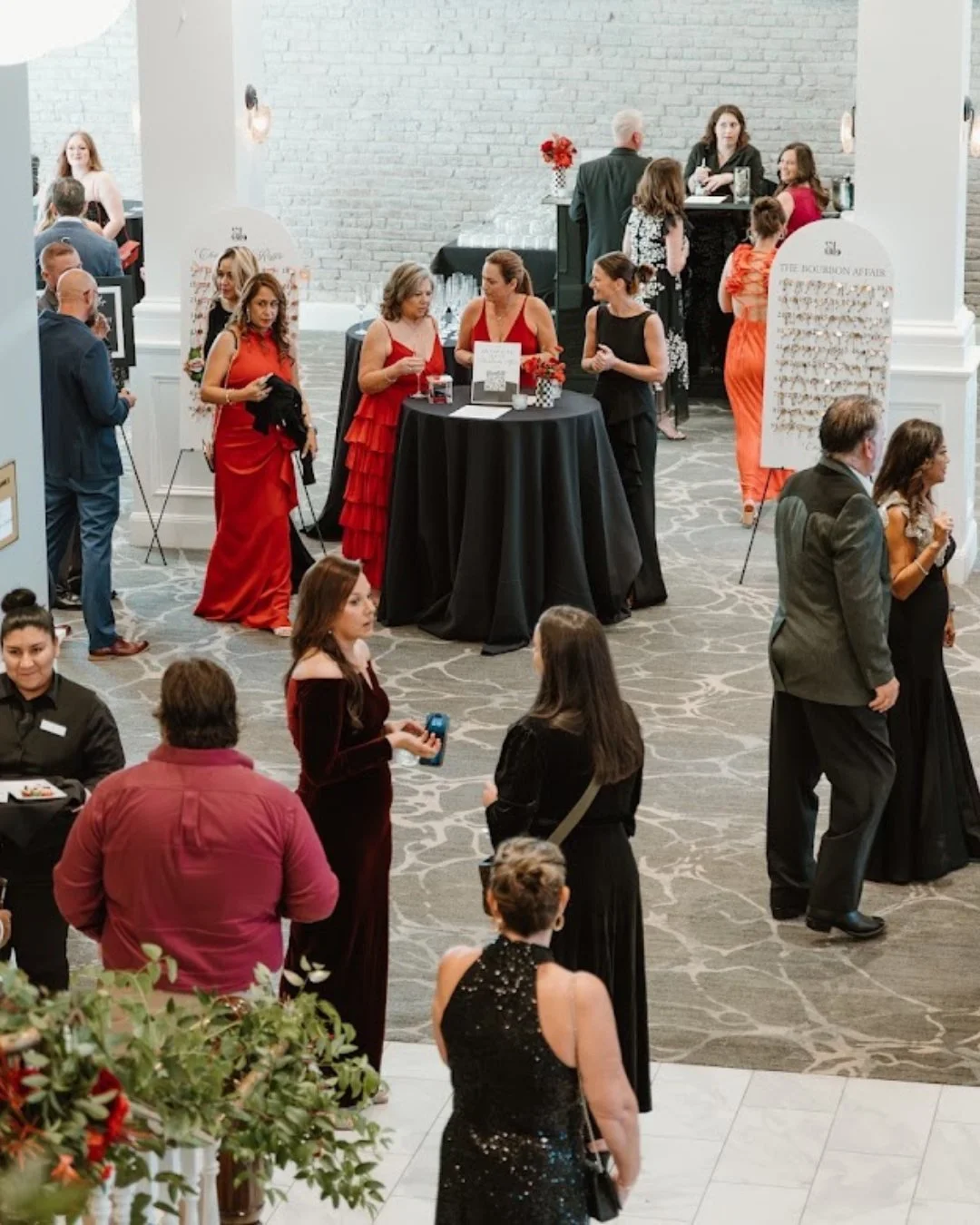 People socializing at an indoor event with a bar area, some in formal attire, including women in red and black dresses and men in suits; a round table with red flowers in the center; signs with jewelry displays; a bar counter at the back with a bartender and customers; a woman with a drink talking to others; the setting appears elegant and festive.