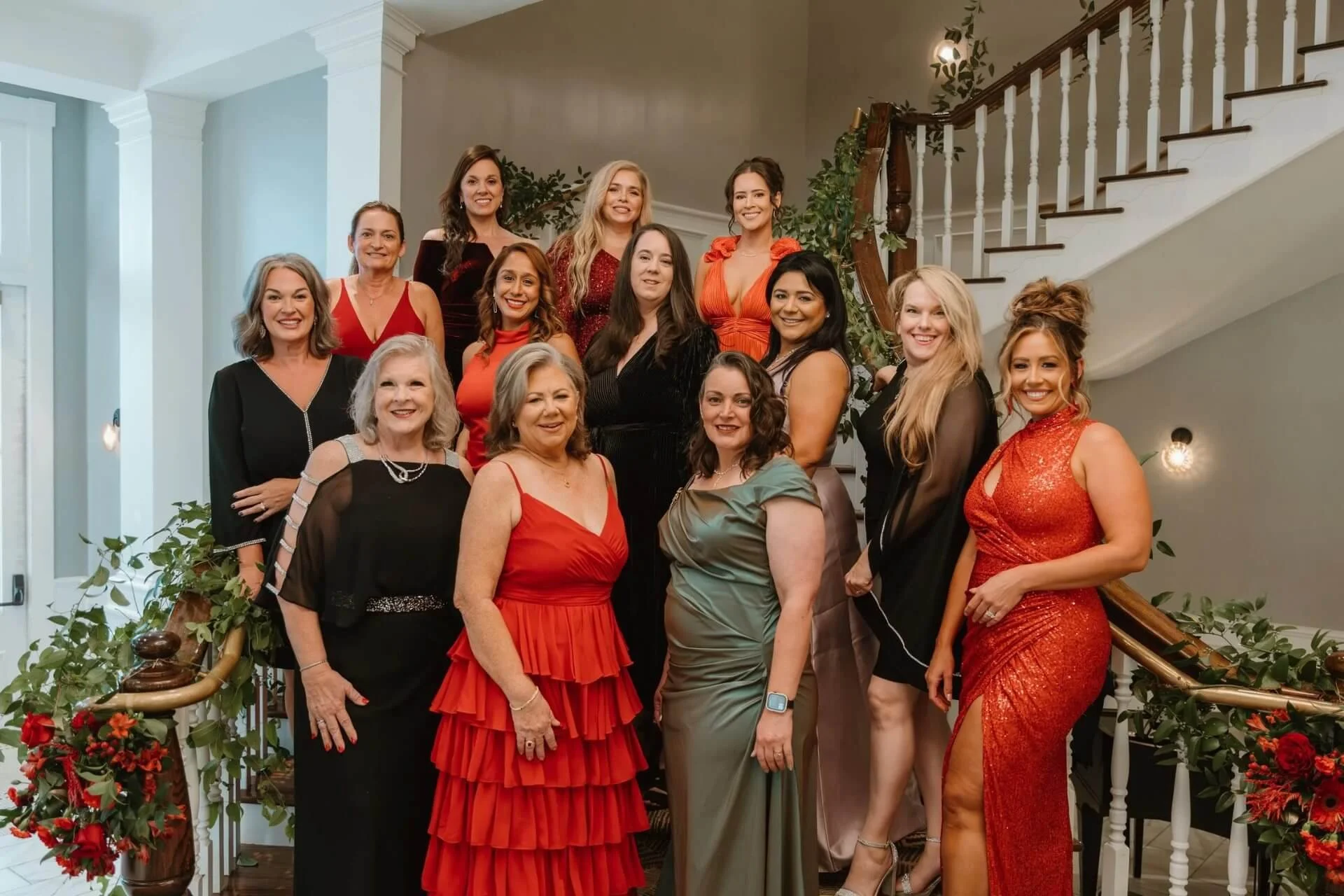 Group of women dressed in formal attire standing on a staircase decorated with greenery and red flowers inside a house.