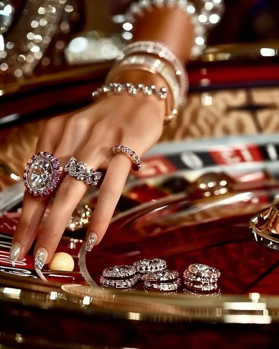 A hand adorned with multiple rings, surrounded by jewelry pieces including rings, bracelets, and earrings, on a reflective surface with a red and black background.