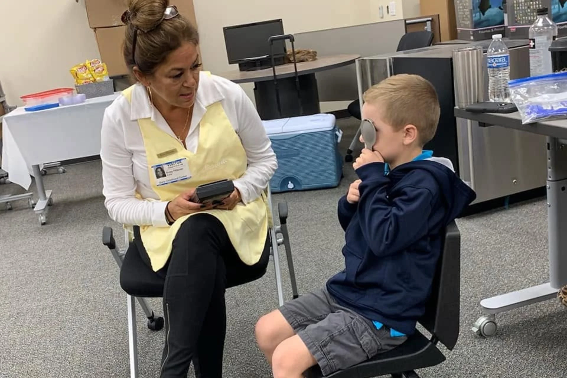 A woman in a yellow apron seated in a chair talking to a young boy in a blue jacket sitting on a chair, in a room with medical supplies and equipment.