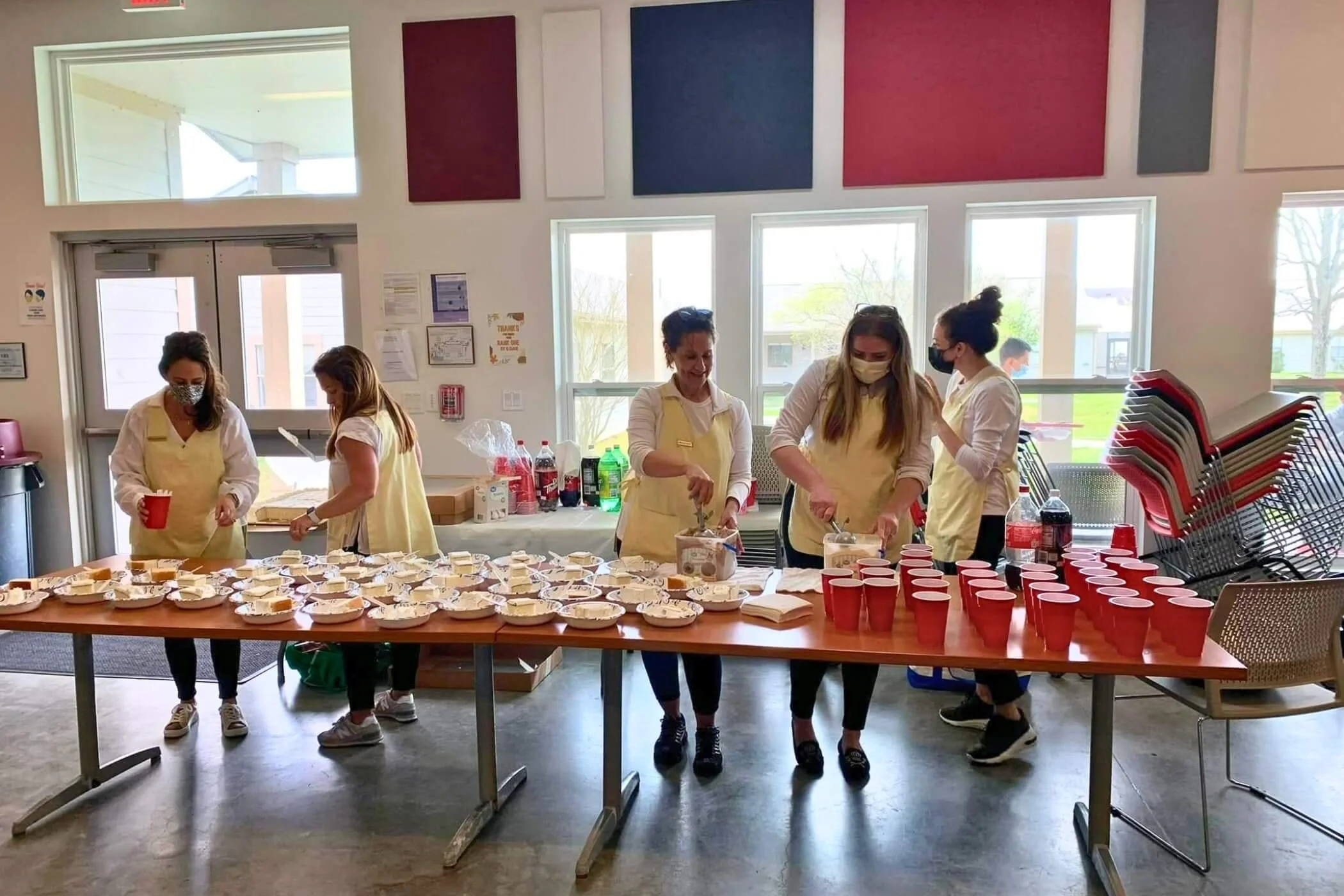 Group of people wearing yellow aprons and face masks preparing food and drinks at a table during a community event in a well-lit room with large windows and colorful wall panels.