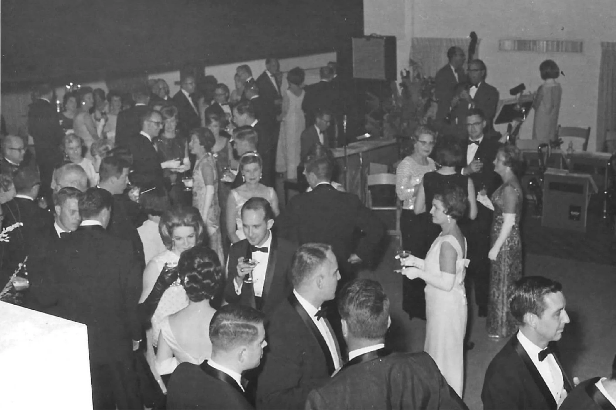 Black and white photo of a social gathering or dance event with men and women dressed in formal attire, including tuxedos and evening dresses, engaging in conversation and holding drinks.