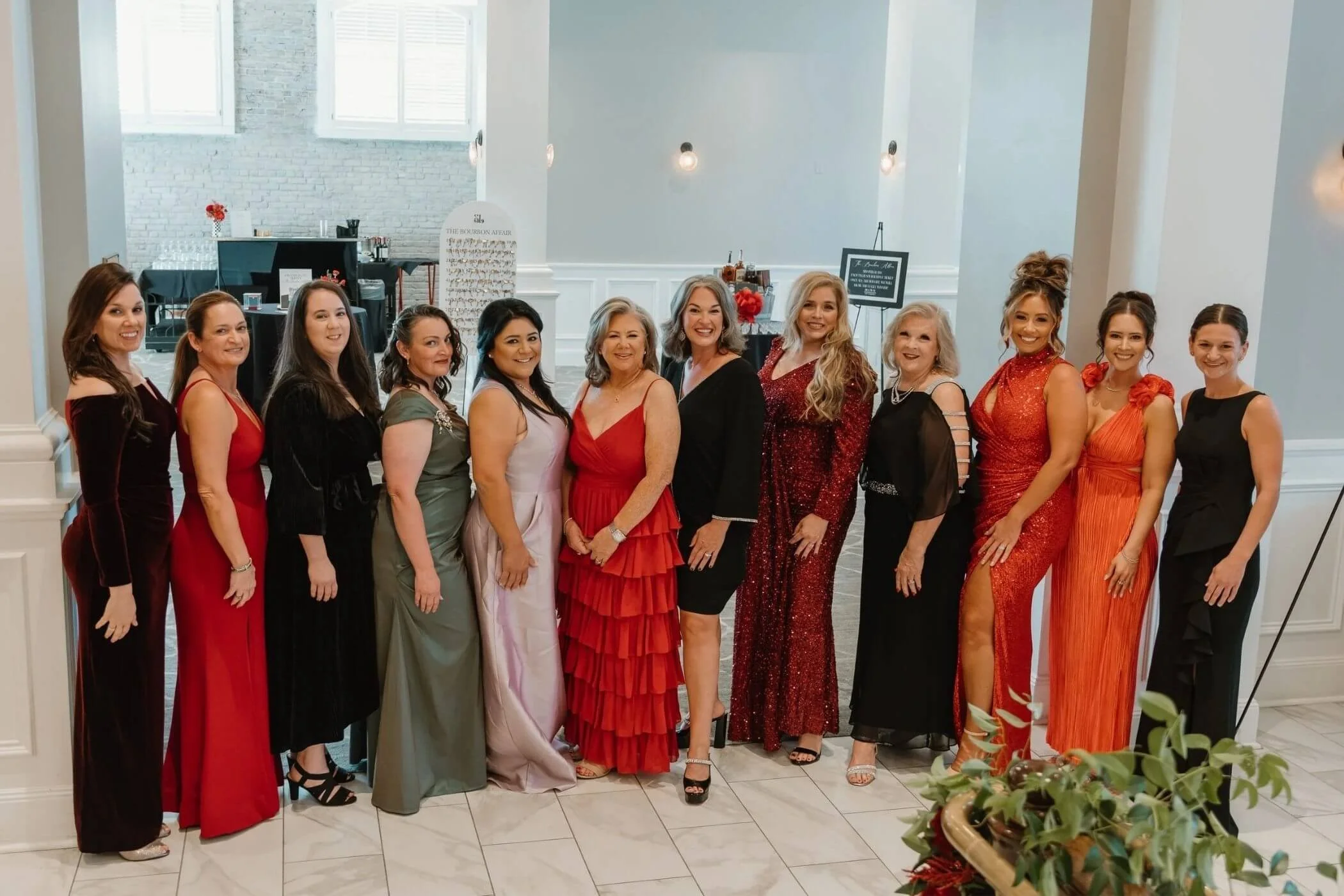 A group of fourteen women dressed elegantly in colorful gowns, standing side by side indoors, in a well-lit room with high ceilings and large windows.