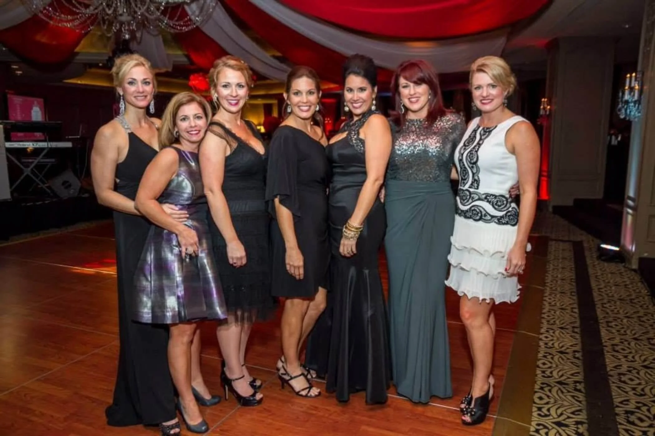 Group of seven women dressed in formal evening wear at a party or event.