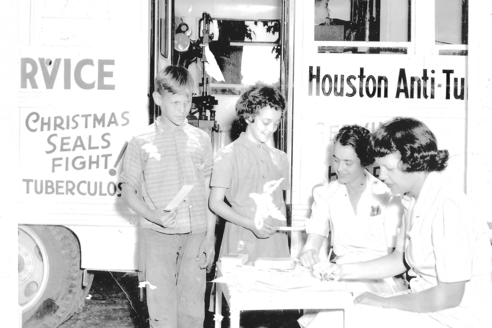 Historically black and white photo of two young students receiving flu shots from two women at a healthcare outreach event in Houston, Texas, promoting anti-tuberculosis measures and Christmas seals.