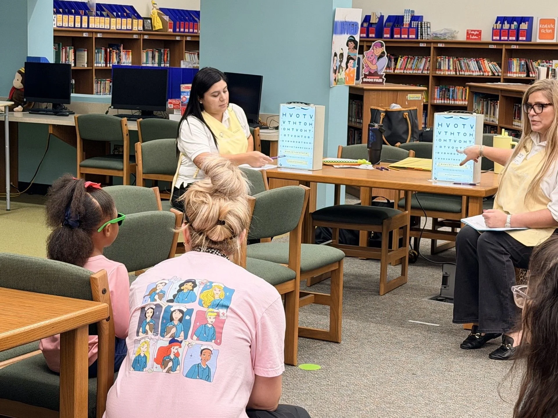 A group of people participating in a literacy or educational program in a library, with two women leading the activity at the front. Children are seated in the foreground, with some wearing pink shirts featuring cartoon characters, and others listening attentively.