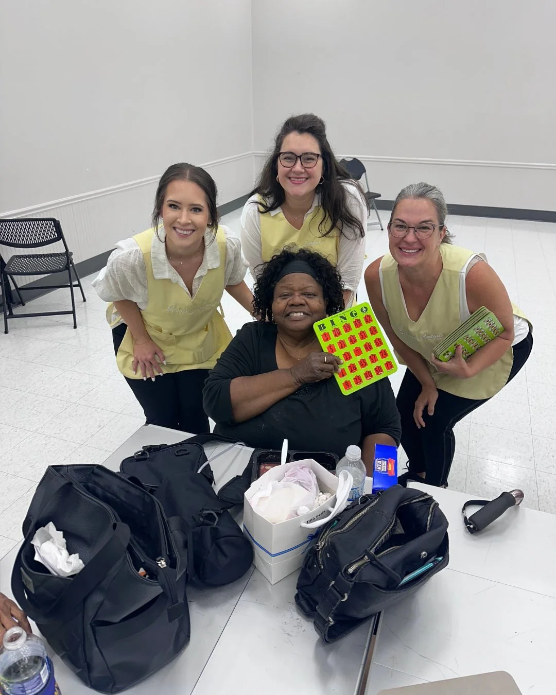 Four women smiling in a room with a table, some bags, and bingo cards, one woman holding a bingo card and another holding a stack of bingo cards.