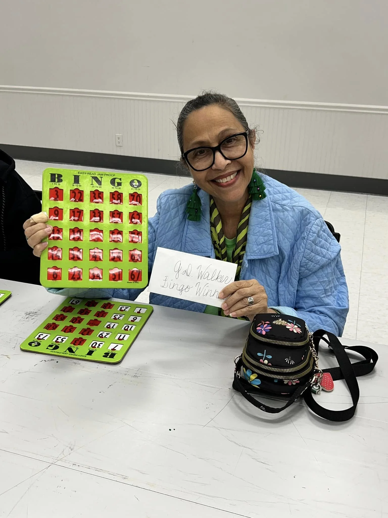Woman with glasses smiling, holding a bingo card, sitting at a table with a second bingo card and a small black floral purse. The background features a plain light-colored wall.