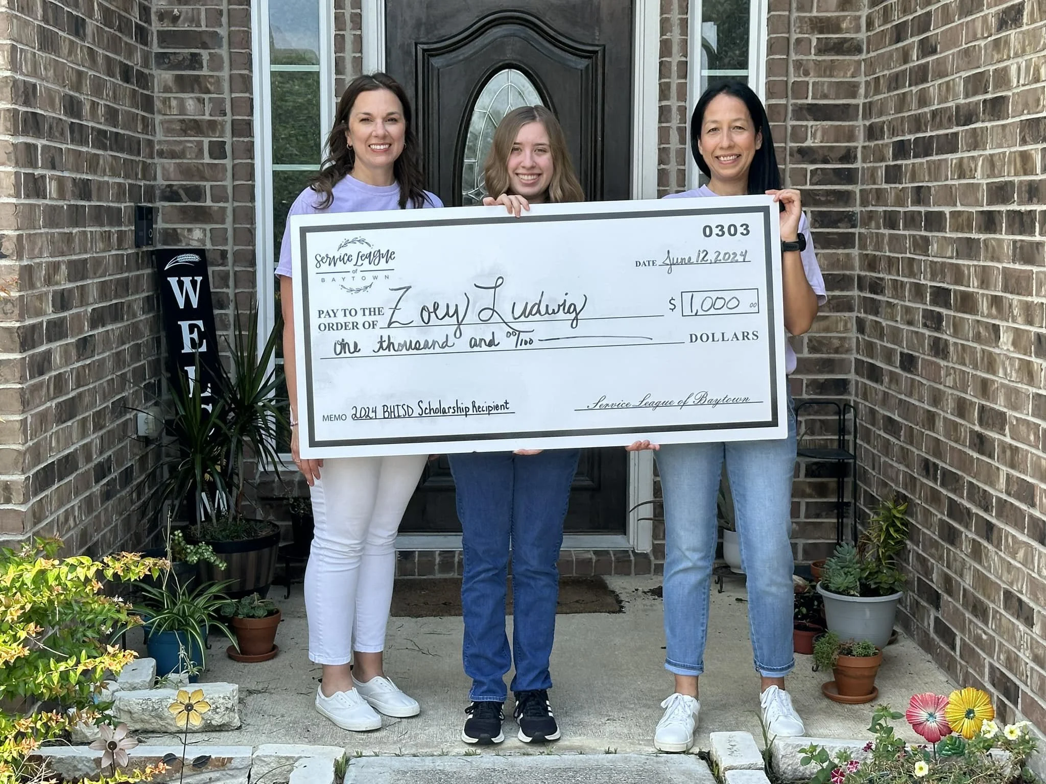 Three women standing in front of a brick house holding a large check, smiling. The check is made out to Zoe Ludwig for $1,000, dated June 12, 2024, from Service League of Baytown.