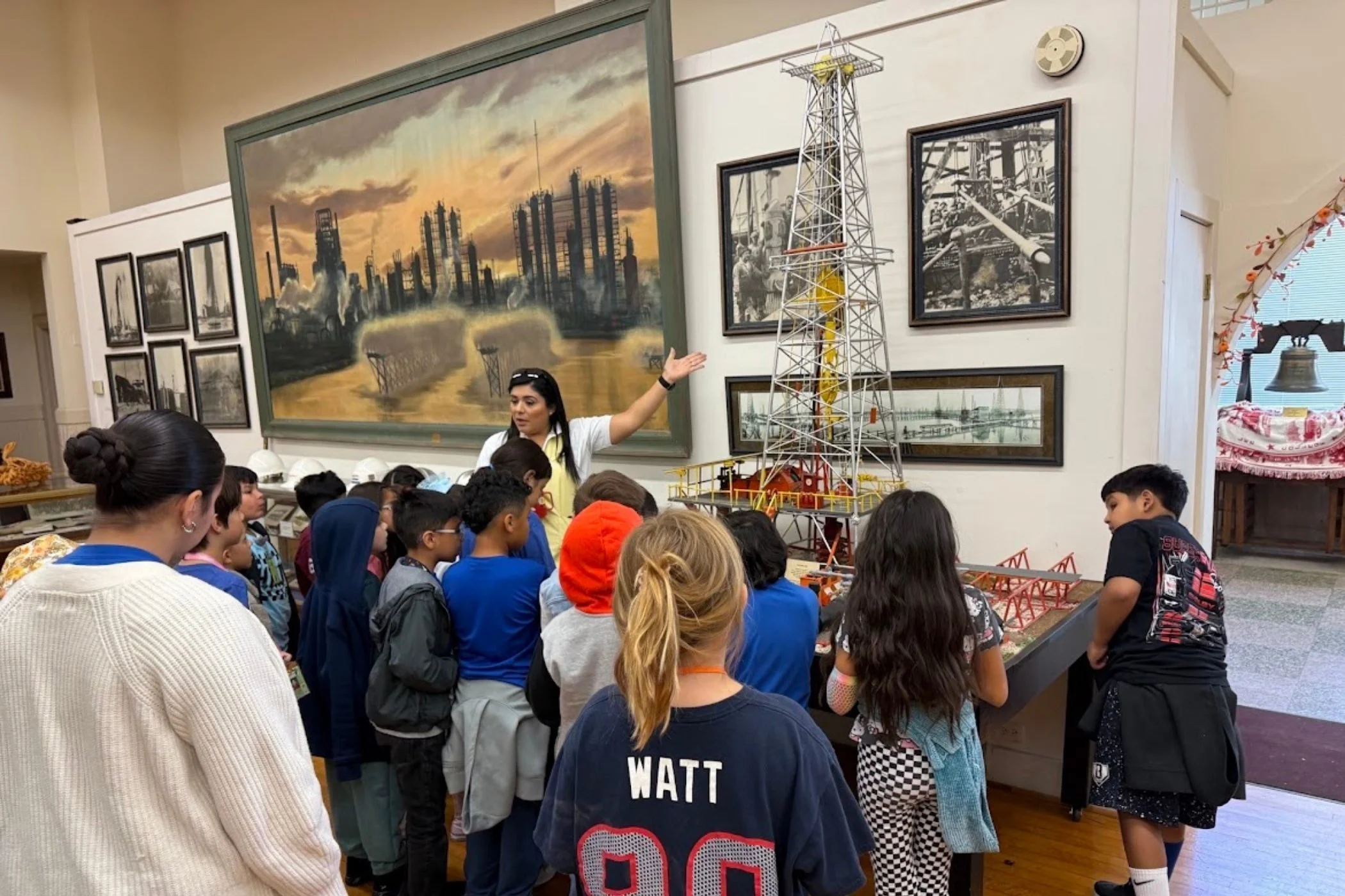 A group of children and adults are gathered in a room looking at a display with models of oil rigs. A woman is speaking and gesturing toward the display. The room has artwork on the walls, including a large painting of an industrial scene with oil rigs and a cloudy sky.