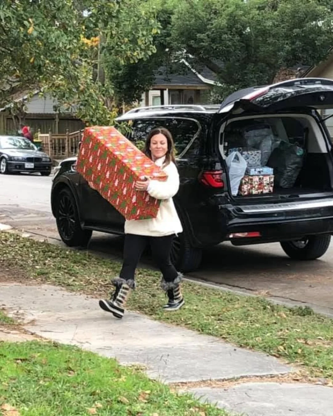A woman carrying a large, wrapped Christmas gift while walking past an open trunk of a black SUV, with holiday-themed presents inside. She is dressed in a white jacket, black pants, and striped boots, and is on a suburban sidewalk with trees and parked cars nearby.