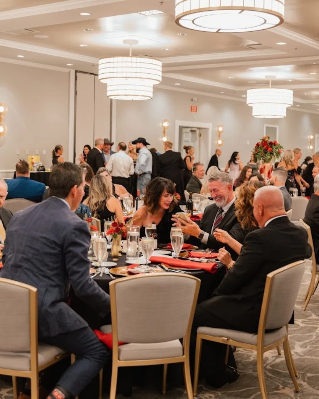 Guests dressed in formal attire enjoying a banquet dinner in an elegant reception hall with large chandeliers and floral centerpieces.