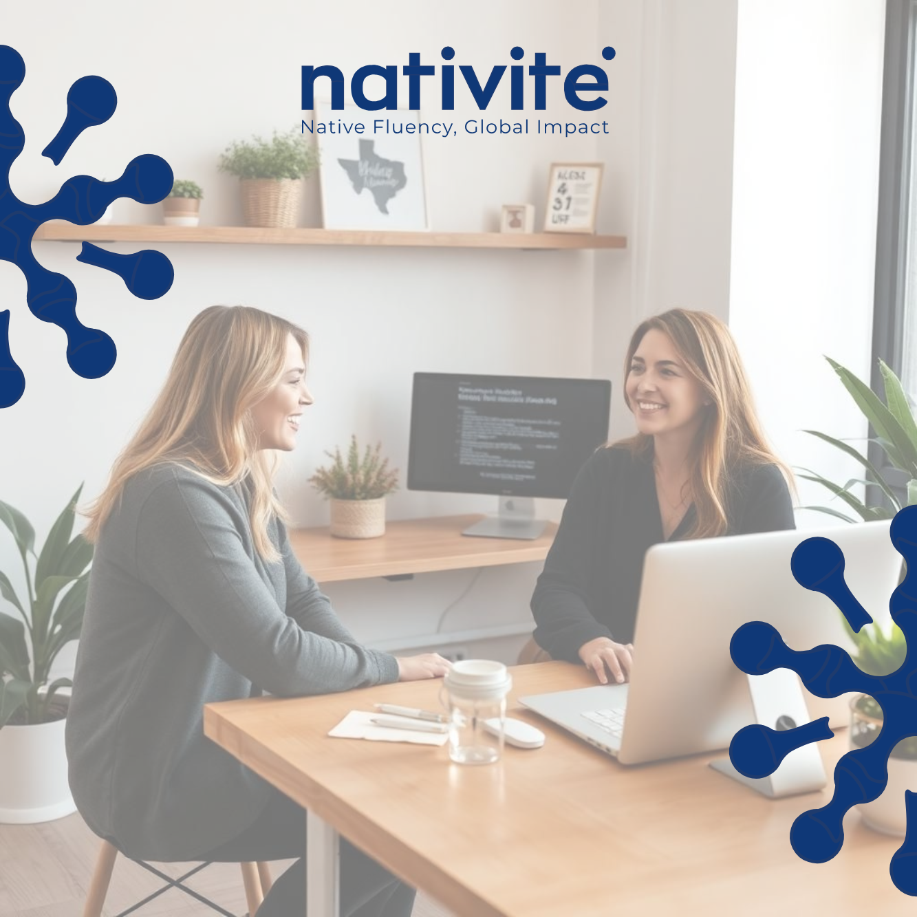 Two women sitting at a wooden table, talking and smiling, in a modern office with plants, a computer, and framed pictures on the wall.