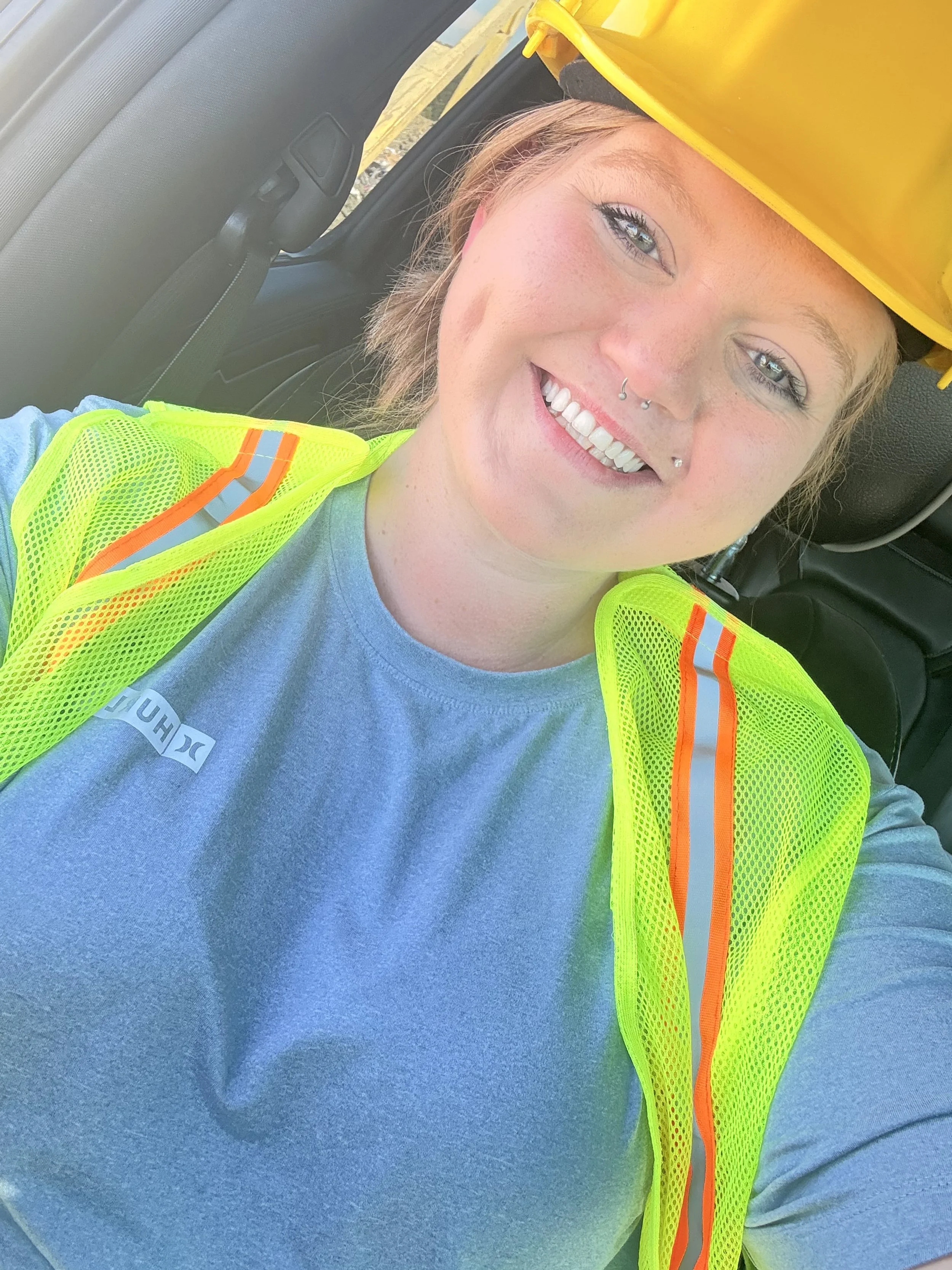 A woman wearing a yellow safety helmet and a bright yellow safety vest is smiling while sitting inside a vehicle. She has freckles, a septum piercing, and multiple nose and lip piercings. Her blue T-shirt has a small logo on it.