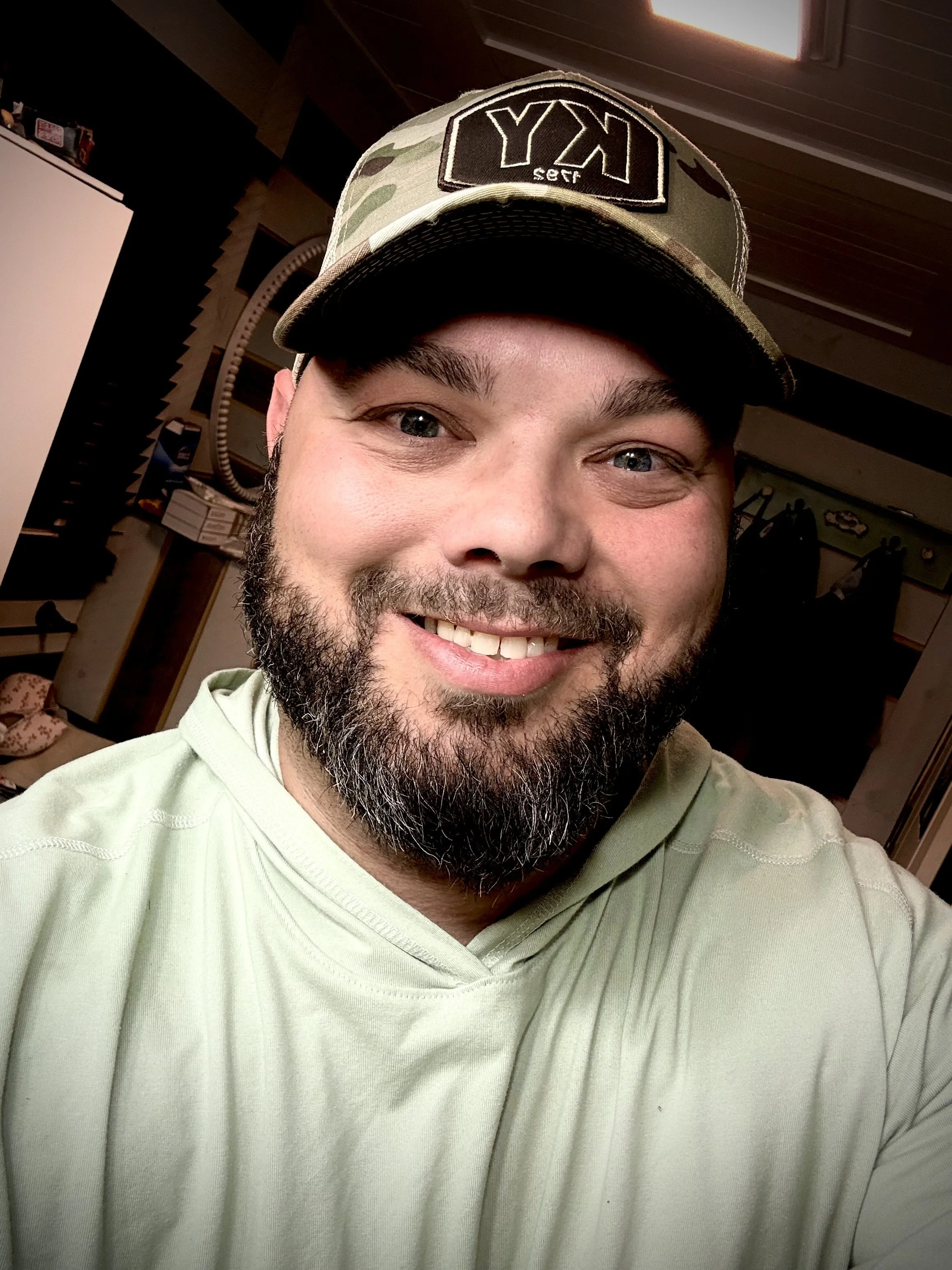 A smiling man with a beard and mustache wearing a green camouflage baseball cap with a patch that says 'XY' and a light green hoodie, inside a room with various objects and wooden walls.