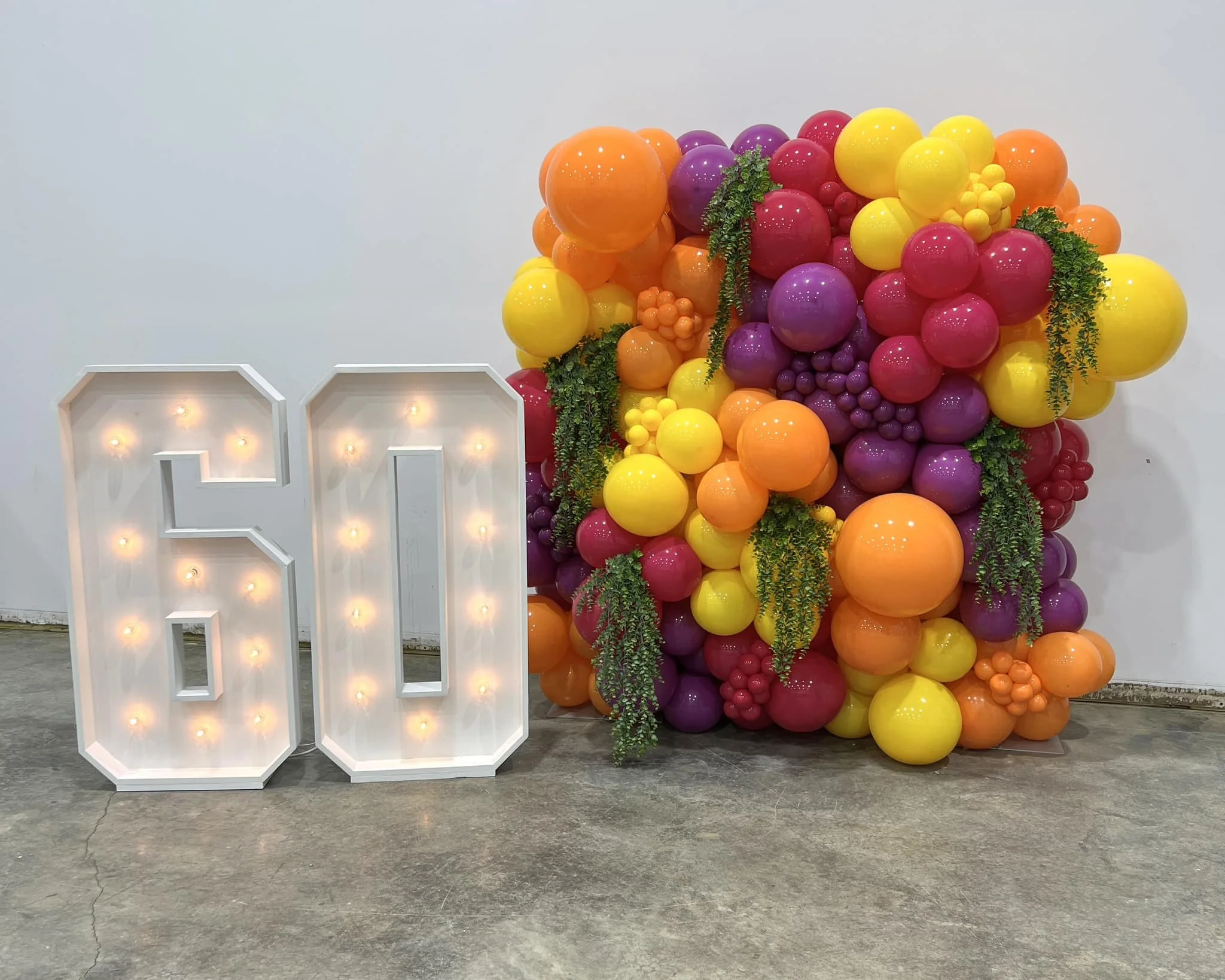 Decorative display featuring a large illuminated number 60 sign beside a colorful balloon arrangement with purple, yellow, orange, and red balloons, accented with green foliage, against a plain white wall.
