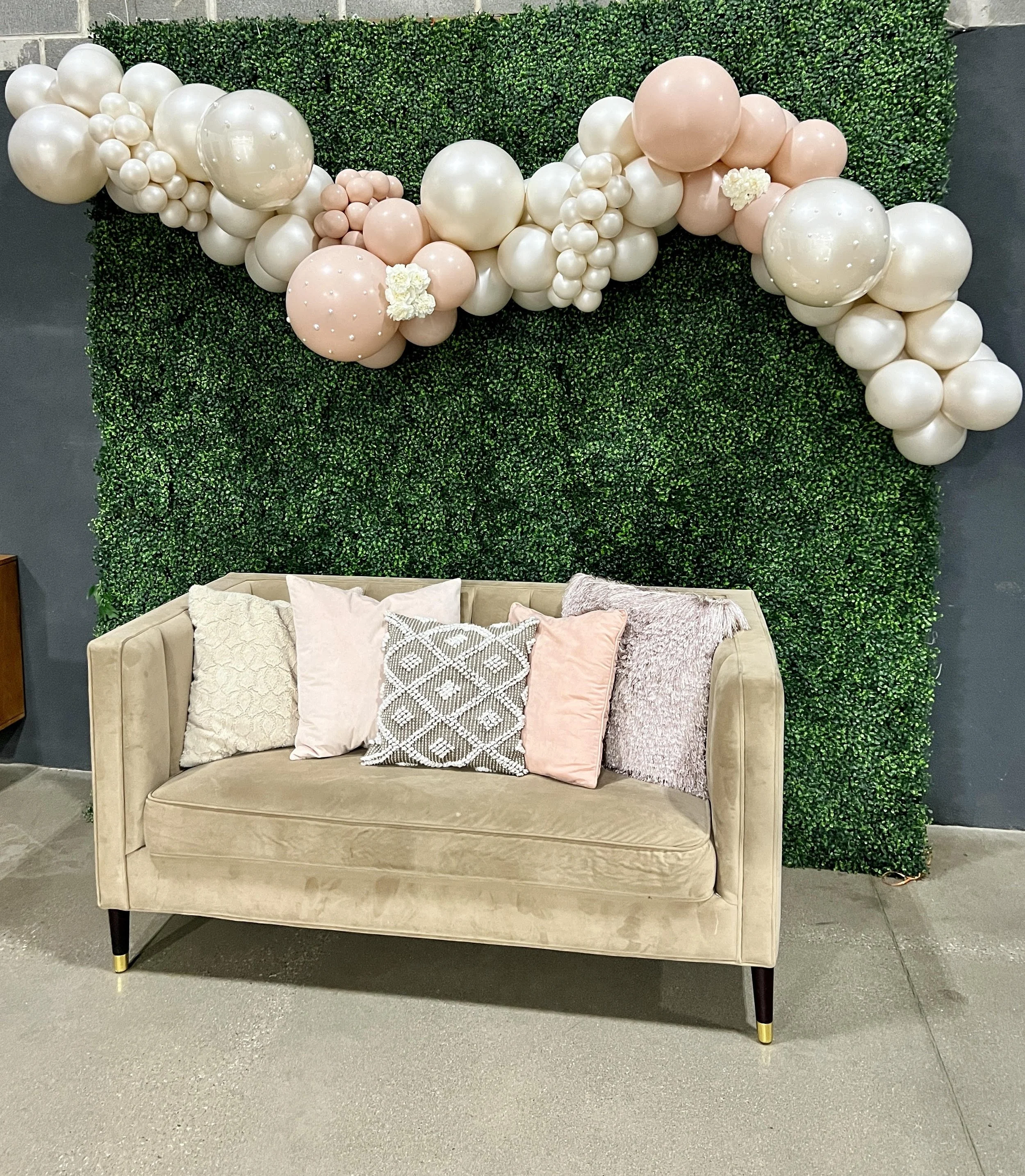 Decorative backdrop with pink and white balloons above a beige sofa with five pillows, set against a green hedge wall.