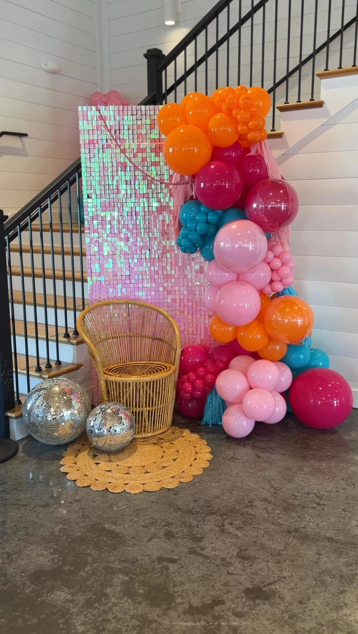 Decorative pink, orange, blue, and red balloons arranged in a cluster against a pink sequin backdrop, with disco balls, a rattan chair, and a crocheted rug in front.