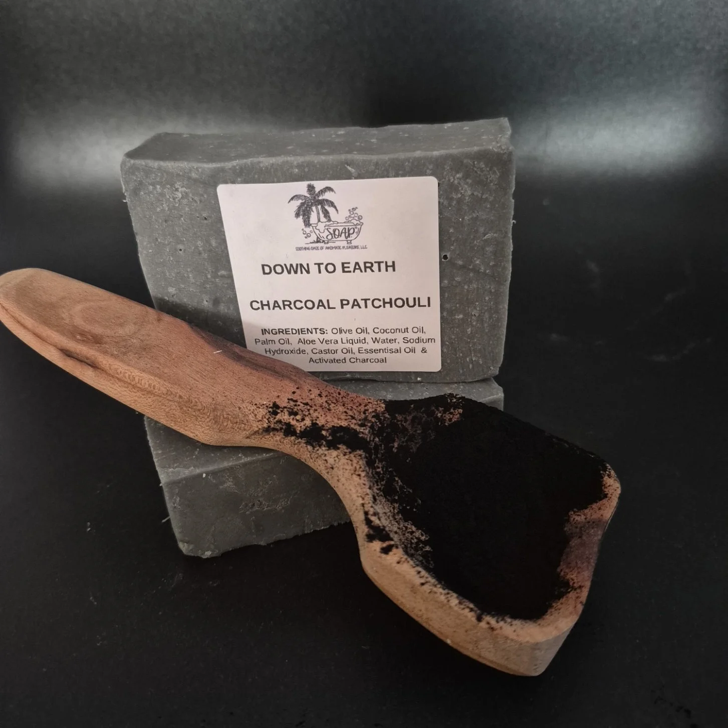 A wooden scoop filled with black activated charcoal powder, placed in front of a grey soap bar labeled 'Down to Earth Charcoal Patchouli' with ingredients listed, against a dark background.