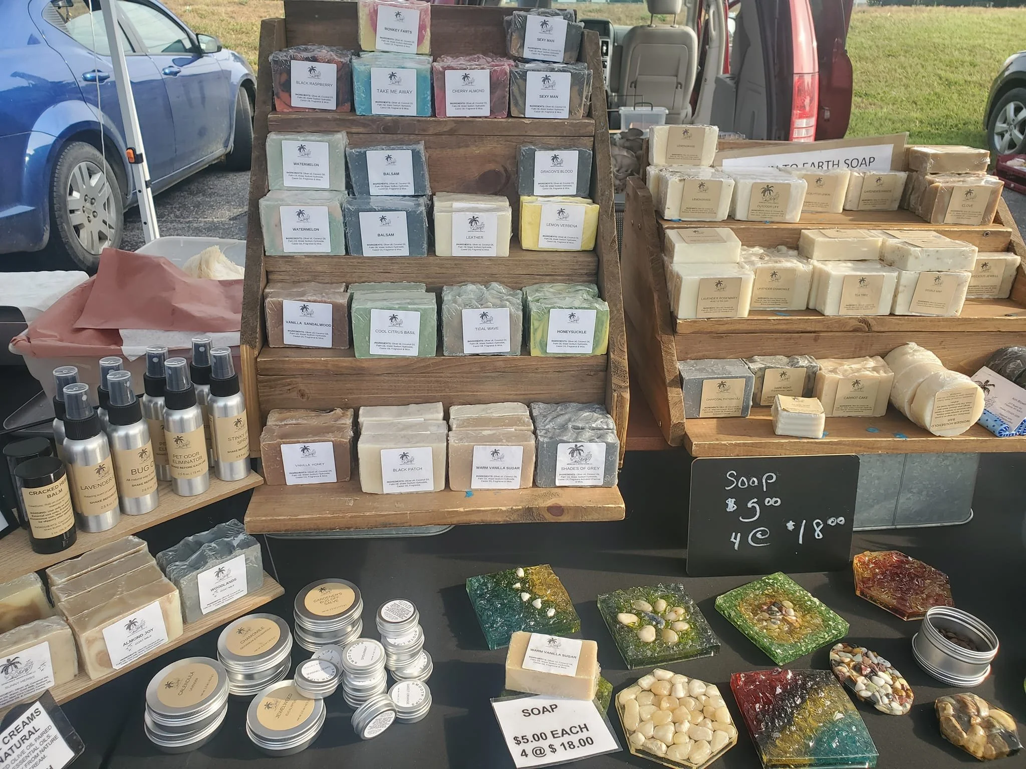 Market stall displaying handmade soap bars, spray bottles, and colorful resin coasters outdoors. Sign indicates soap costs $5 each or 4 for $18.