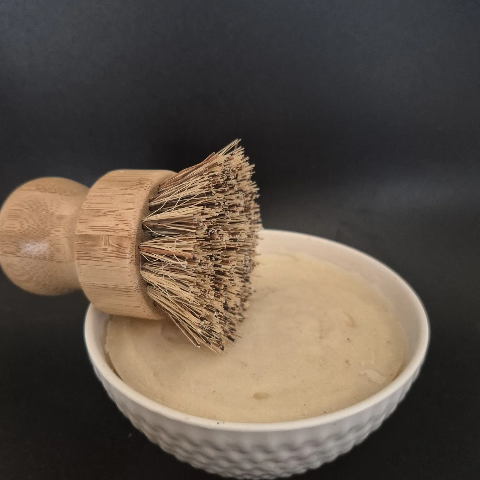 A wooden dish brush resting on a bowl of soap or cream against a black background.