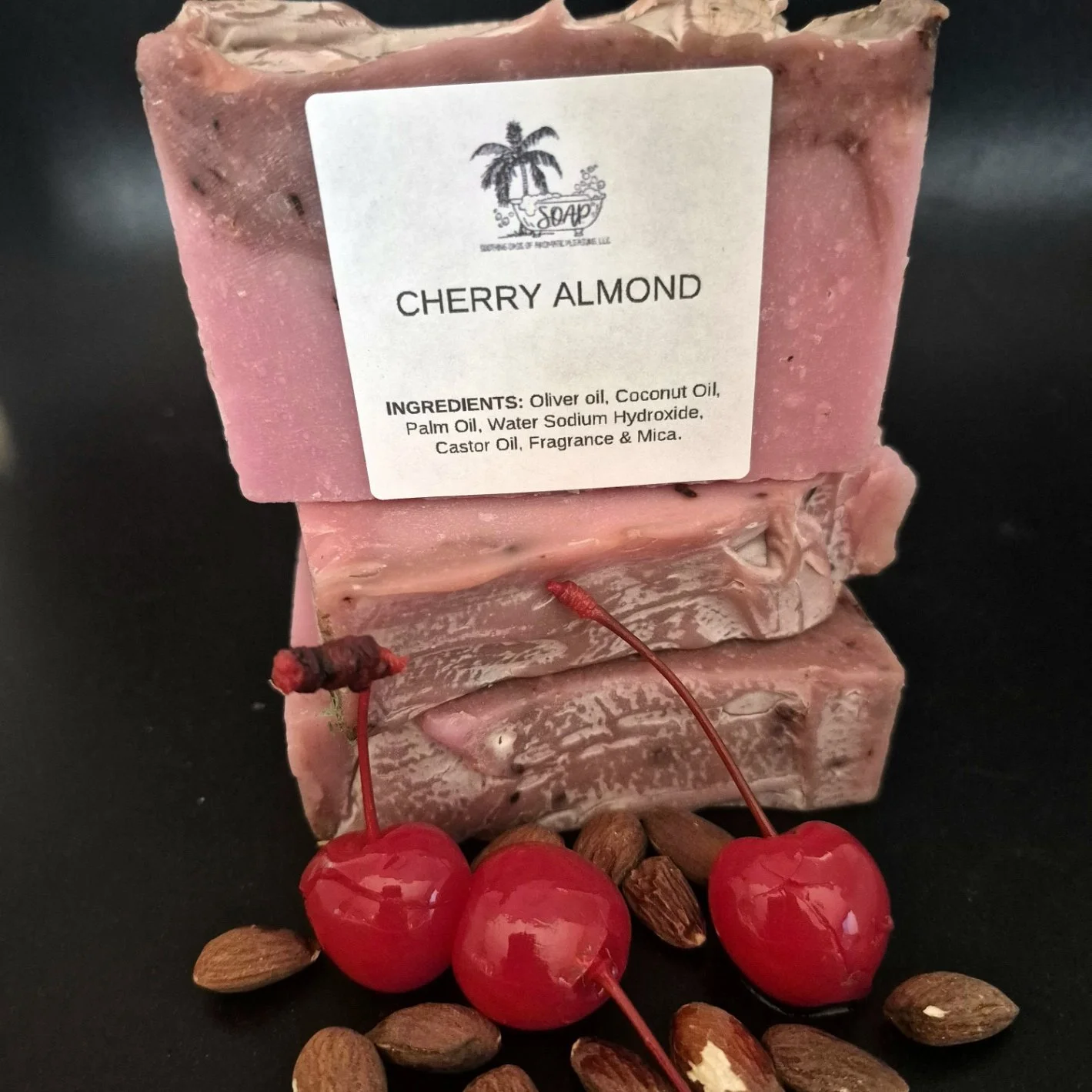 A bar of Cherry Almond soap with a label showing ingredients, placed on a black surface surrounded by cherries and almonds.