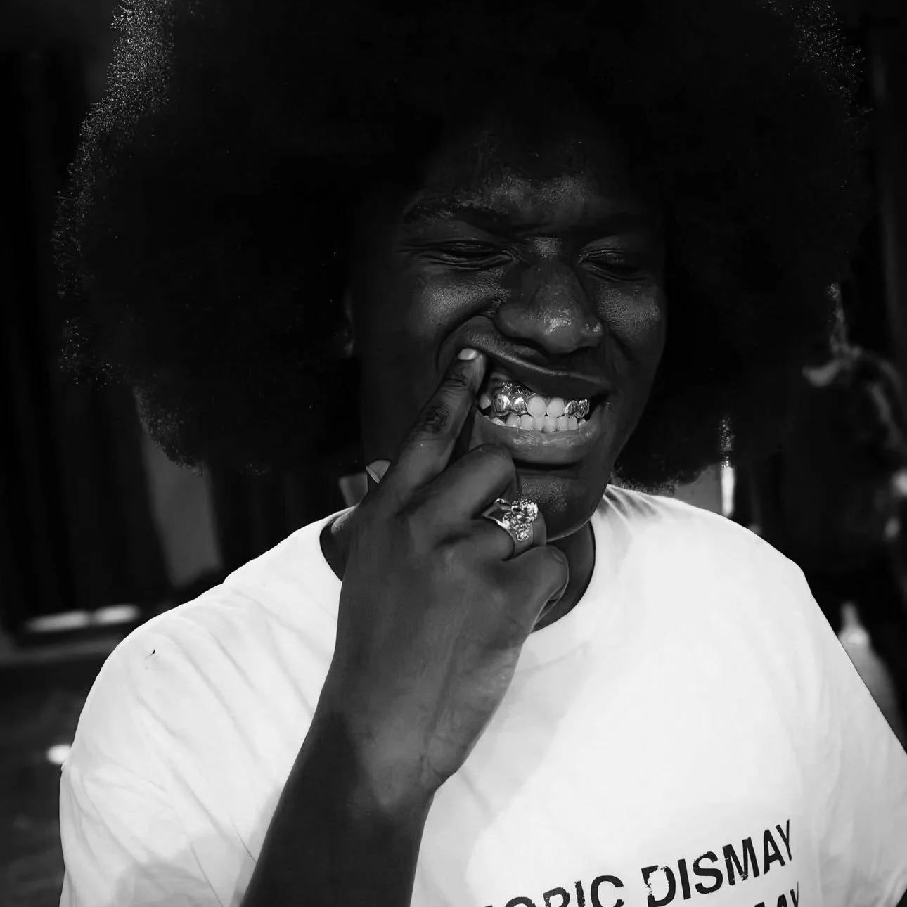 A woman with large natural hair making a playful face, pinching her nose with her finger, smiling showing her teeth and wearing a ring and a white T-shirt.