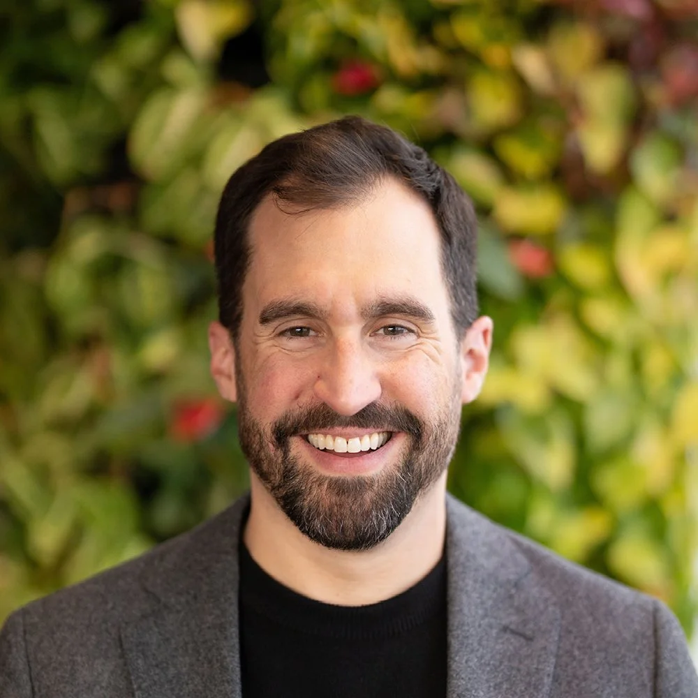 A smiling man with dark hair, a beard, dressed in a black shirt and gray blazer, standing in front of a leafy background.