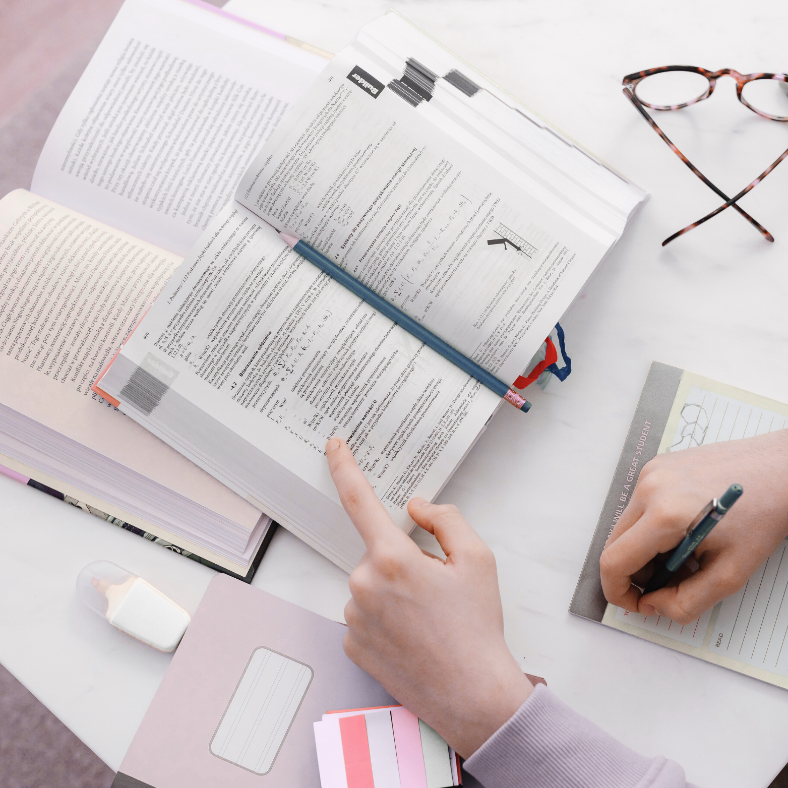 Person studying with open textbooks, a pair of glasses, notebooks, and writing tools on a white desk.