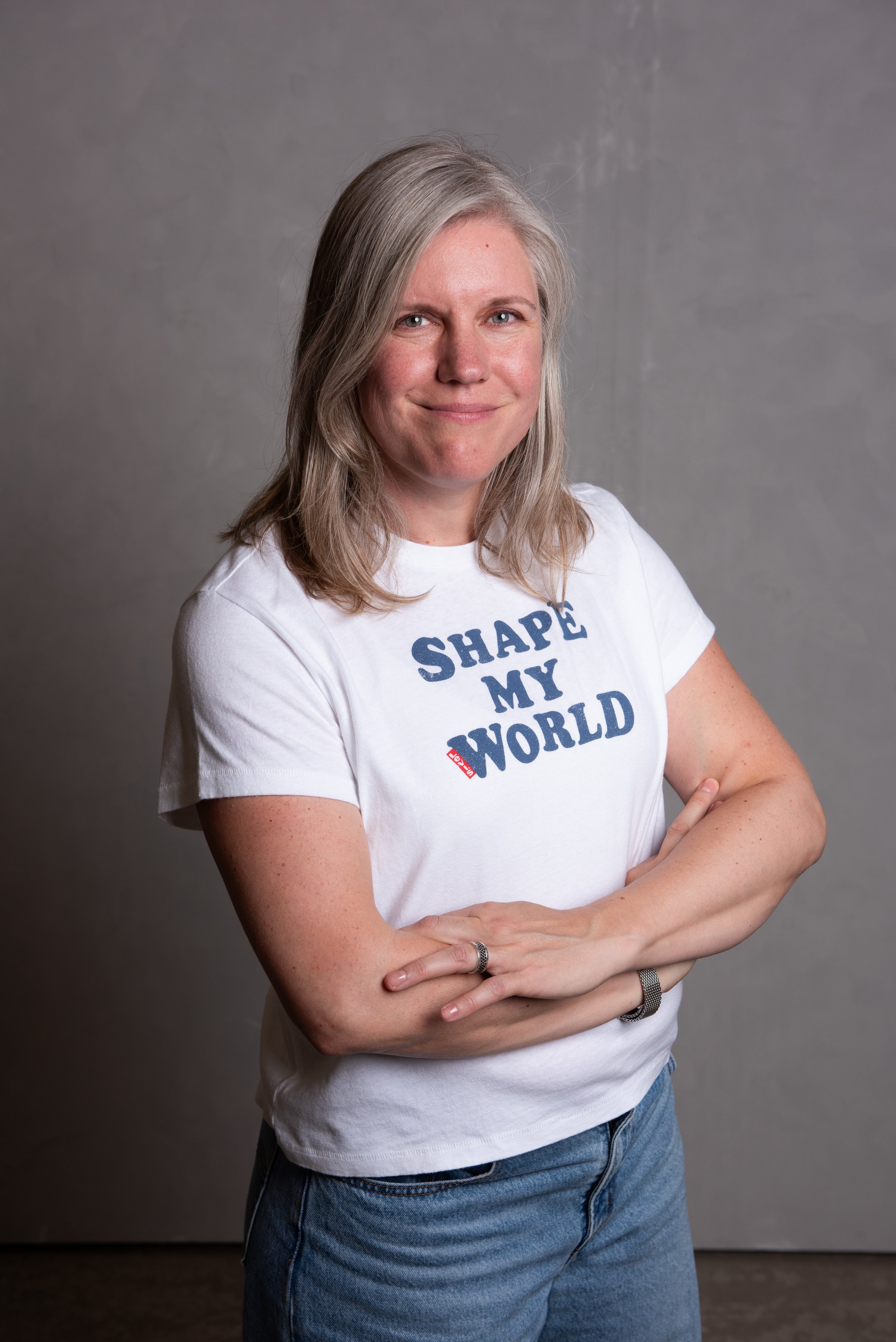 A woman with shoulder-length gray hair, smiling, wearing a white t-shirt that says 'Shape My World' and blue jeans, standing with her arms crossed against a plain gray background.