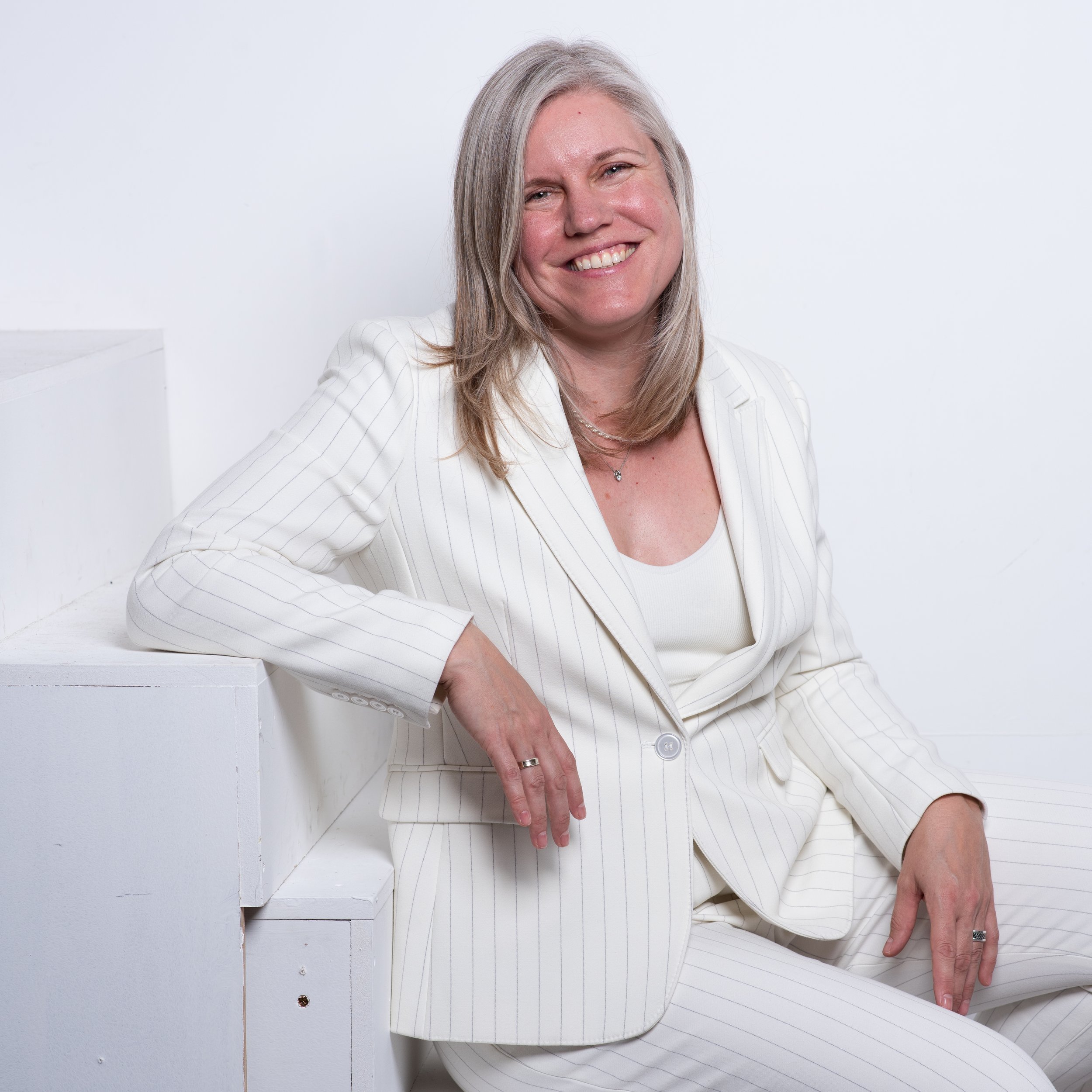 A woman with shoulder-length gray hair, smiling, wearing a white pinstripe suit and sitting on white steps against a plain white background.