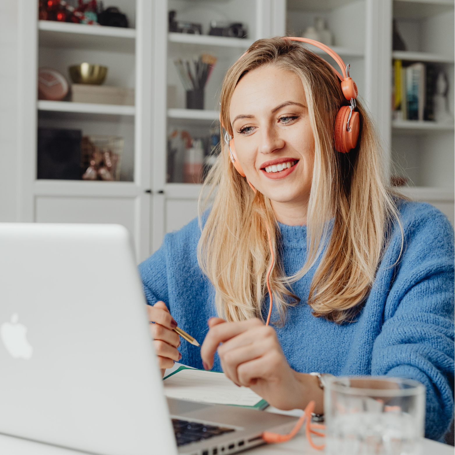 A woman with blonde hair wearing a blue sweater and pink headphones smiling while working at her laptop in a home office.