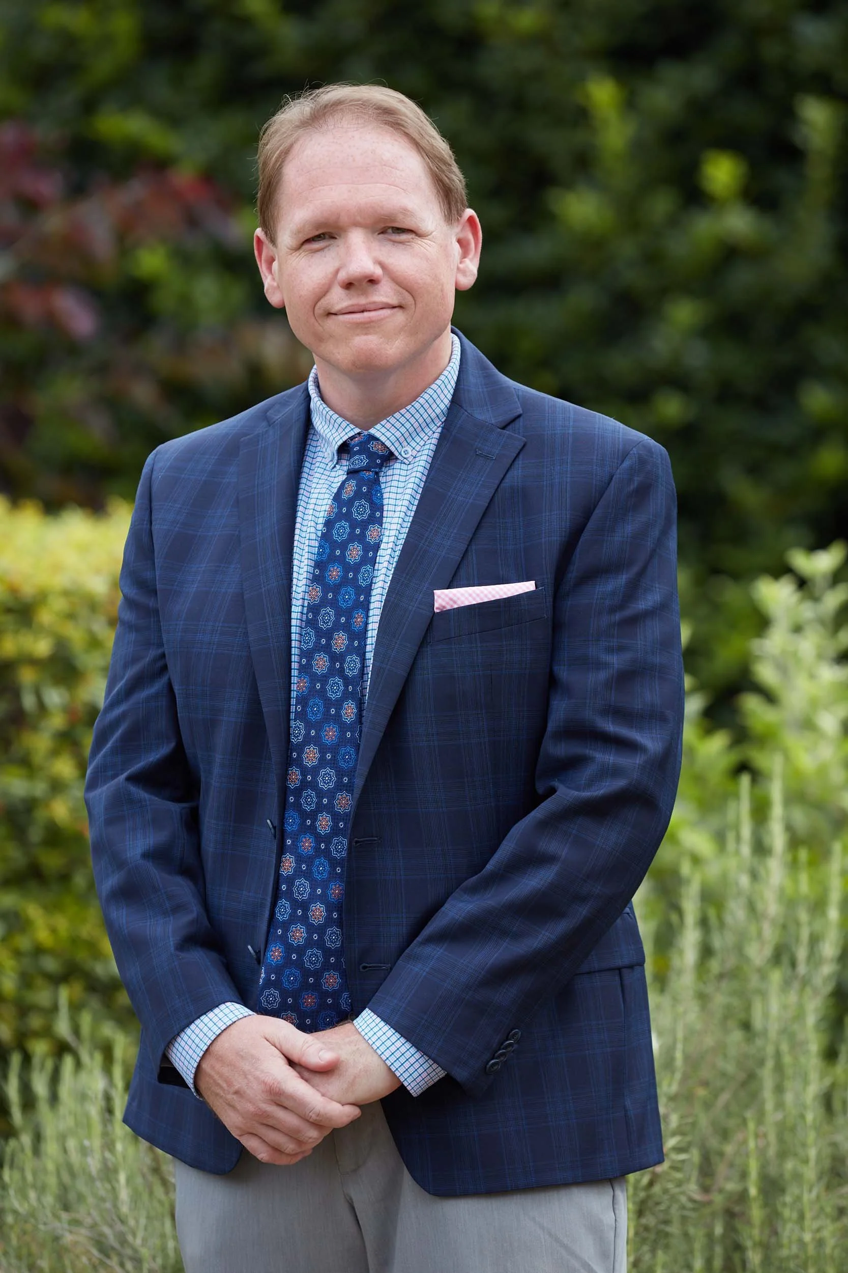 A man in a blue checkered suit and a patterned blue tie standing outdoors in front of greenery.