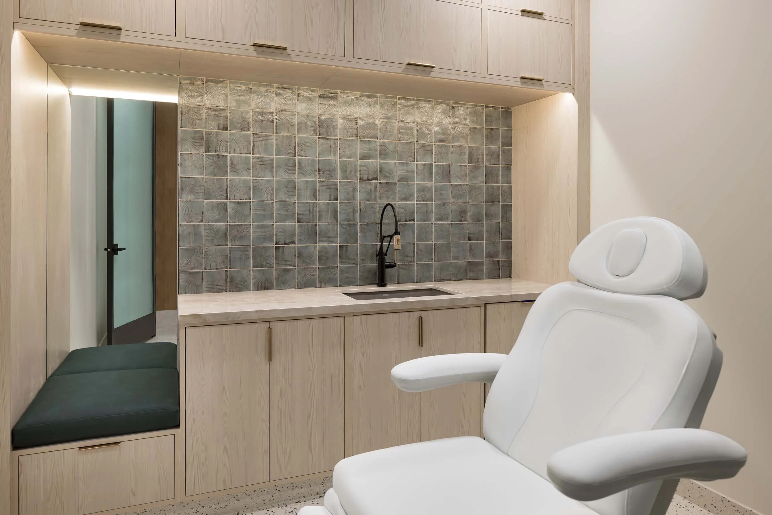 Dental examination room with a white dental chair, light wood cabinets, a small sink with black faucet, and gray tiled backsplash.