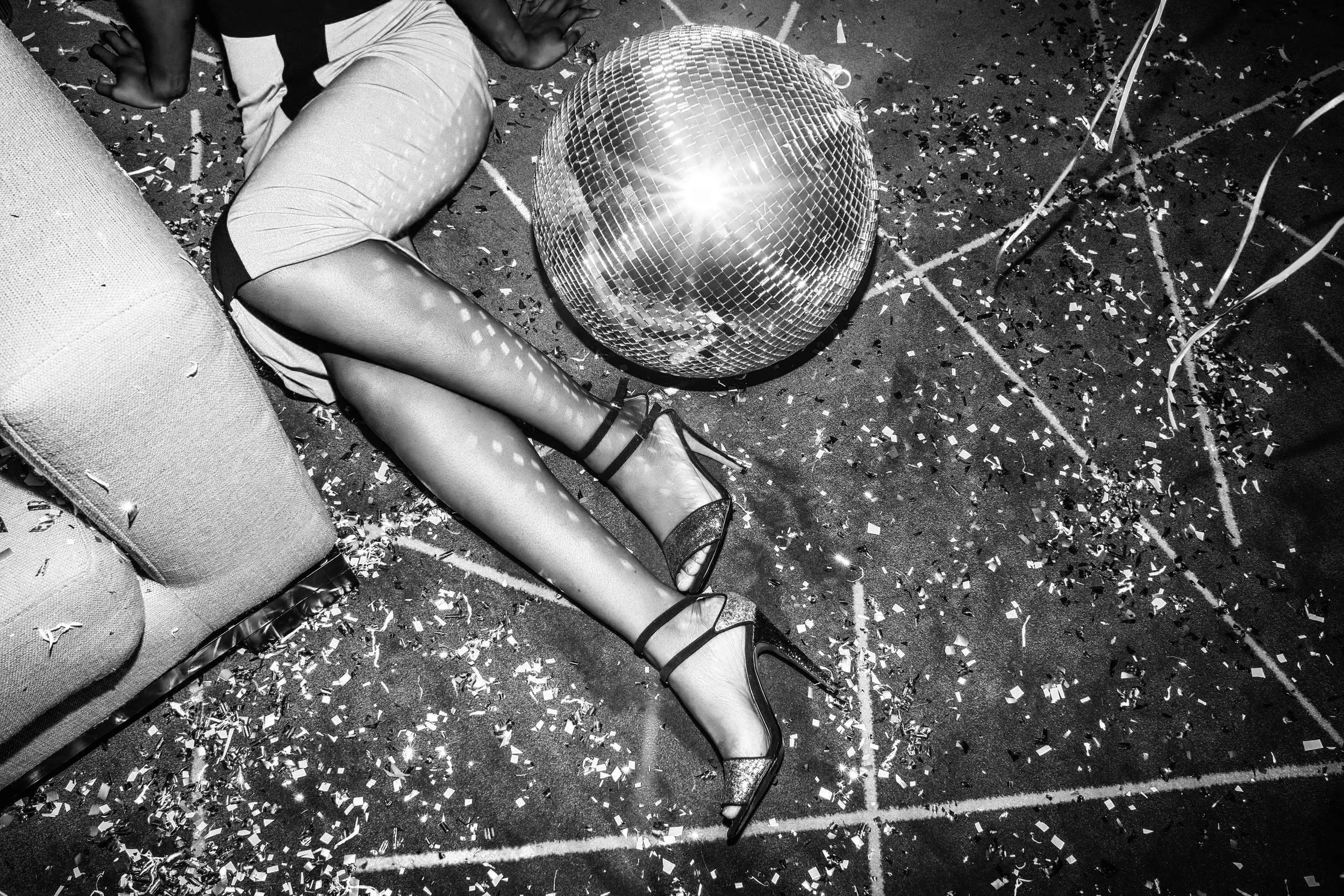A person lying on the floor next to a disco ball, surrounded by confetti and party decorations, wearing high heels and semi-transparent stockings.